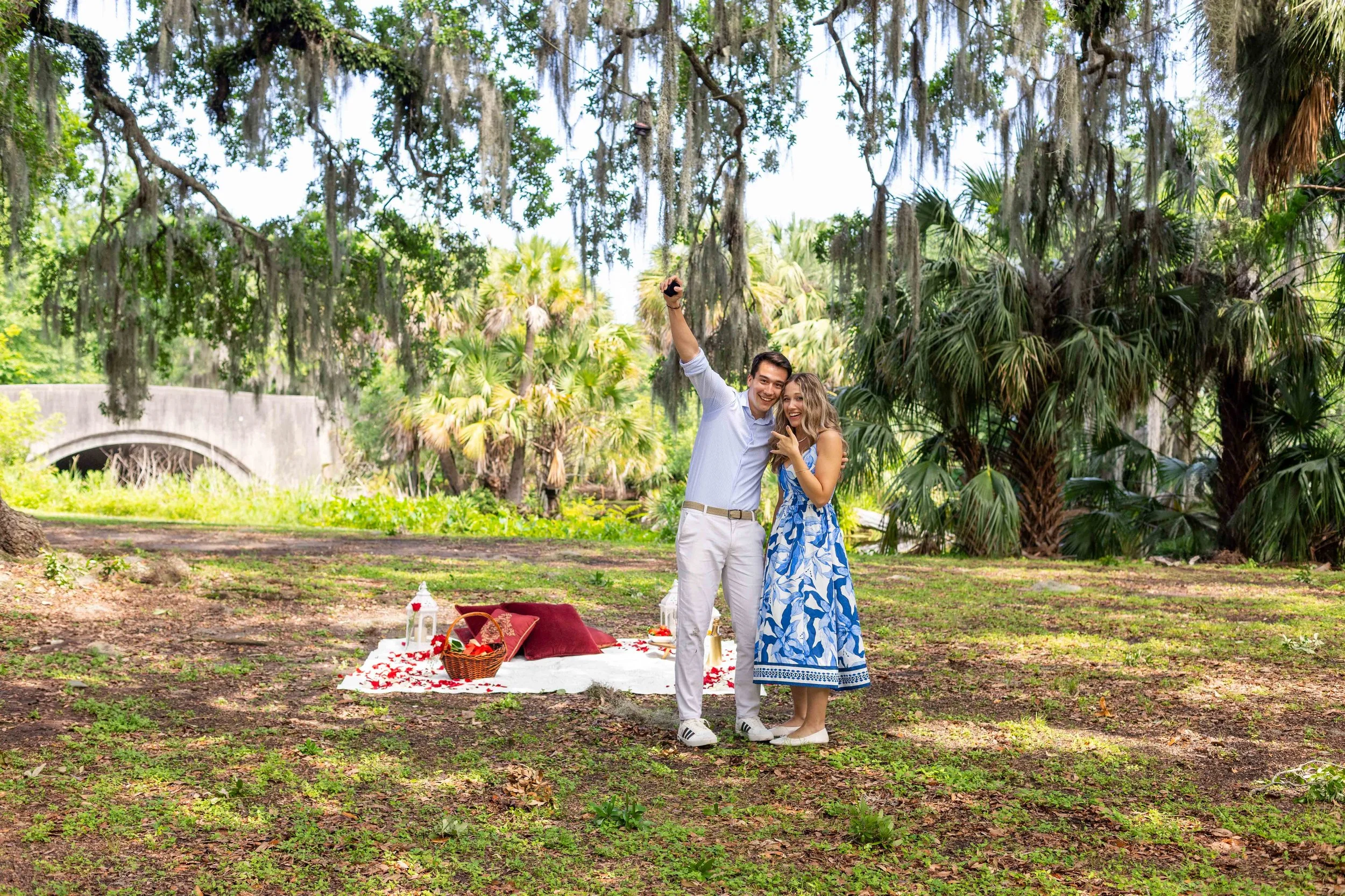 A man fist pumps next to his surprised girlfriend in front of a surprise proposal picnic in New Orleans City Park.