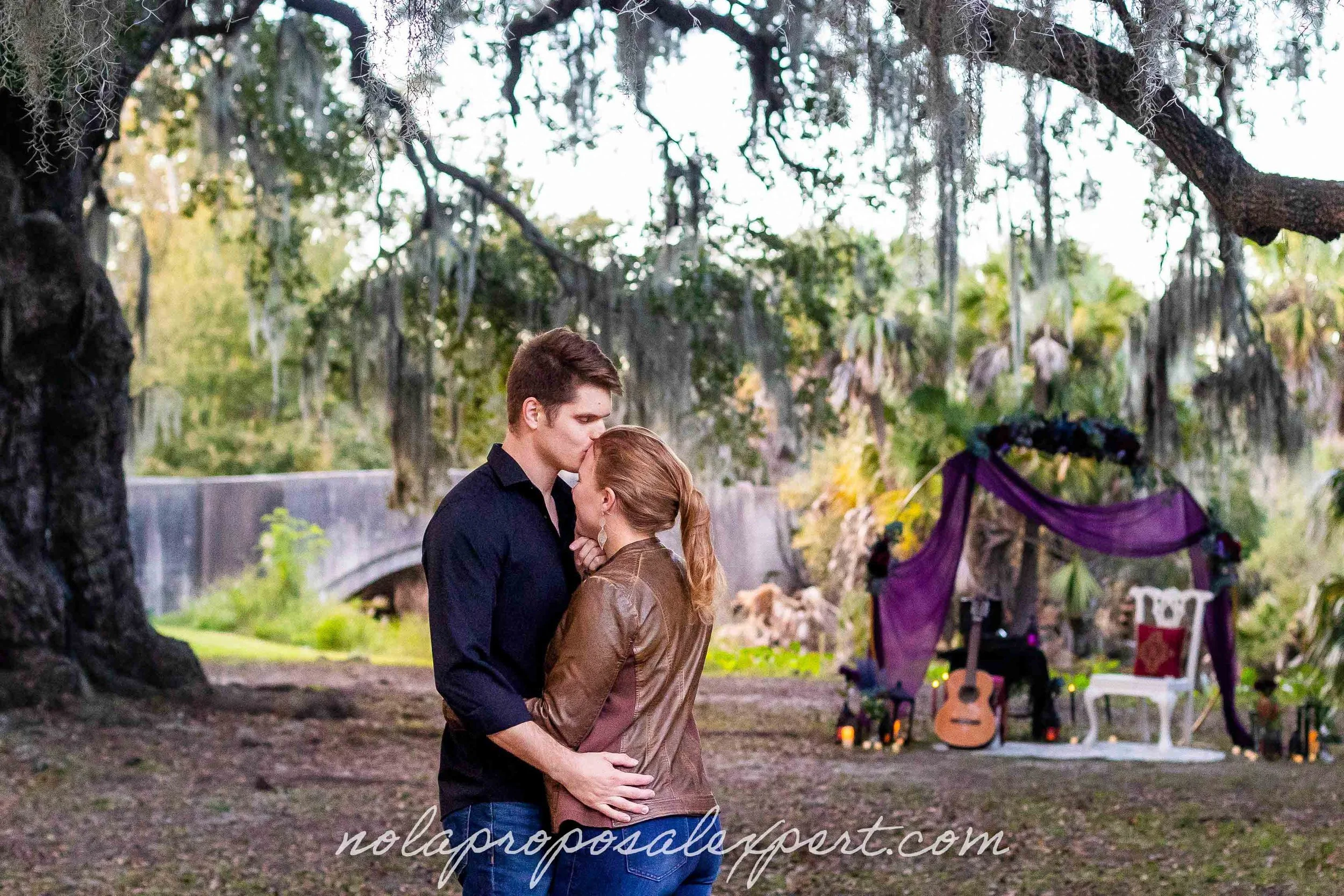 A man kisses his fiance's forehead in front of a beautiful proposal picnic setup under an Oak in New Orleans city park .