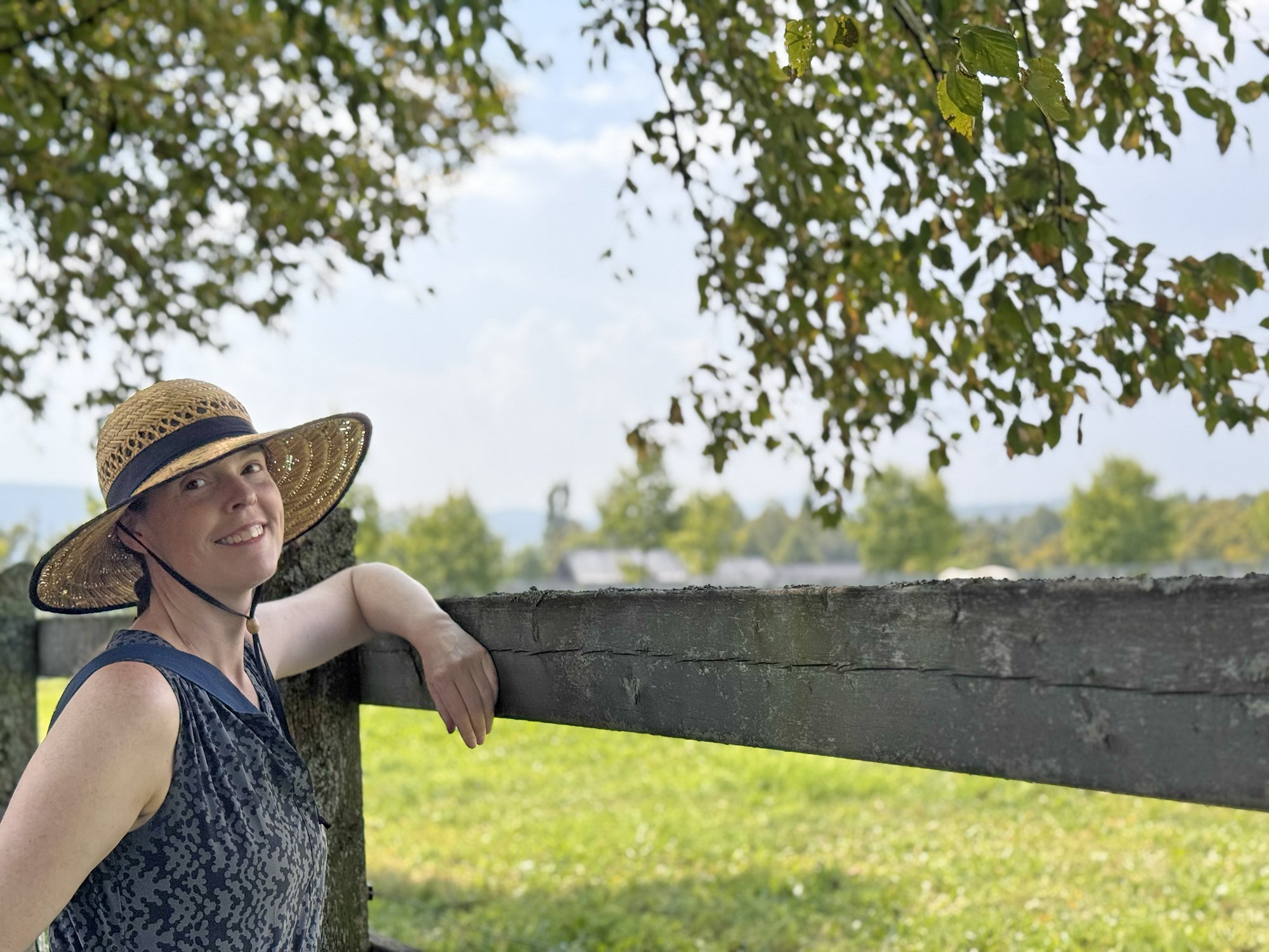 Dana Wheeles wearing a straw hat and leaning against a fence by a grassy field.