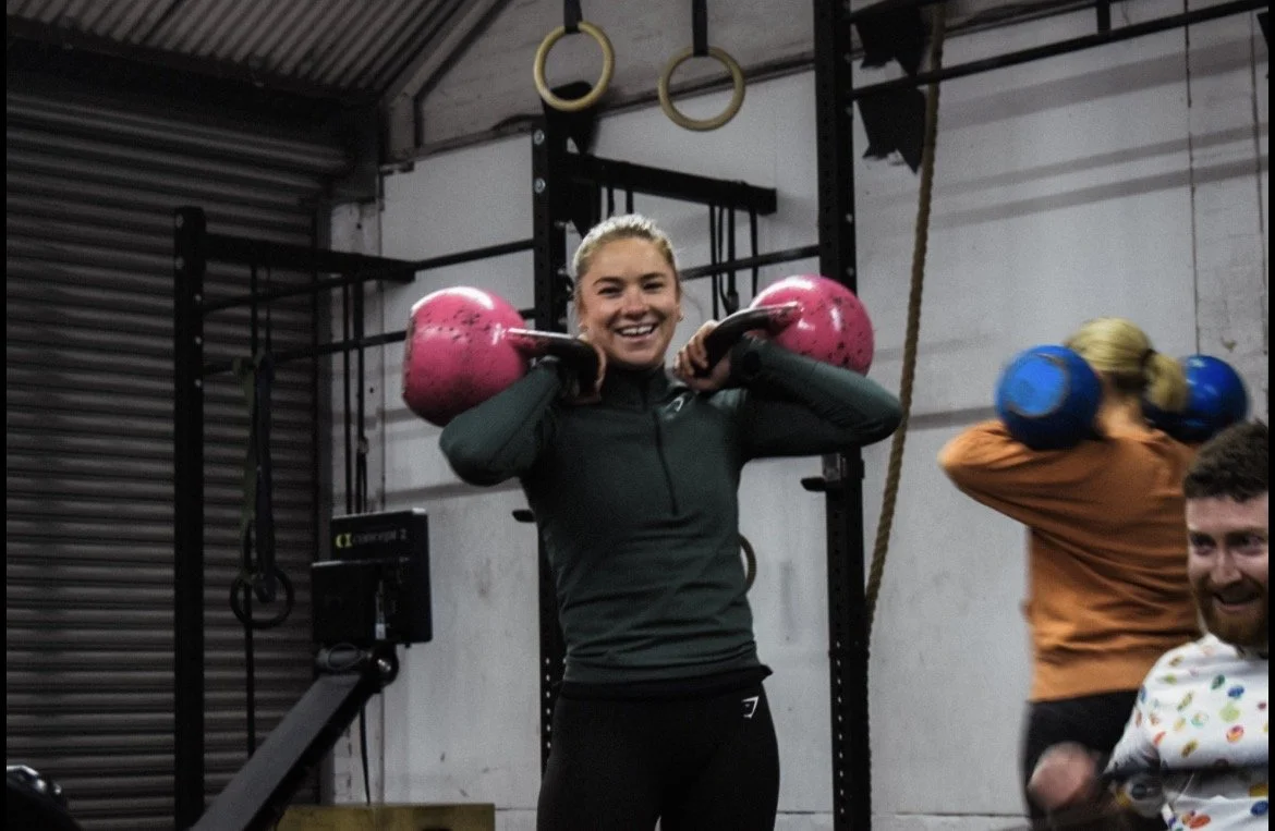Image of member smiling mid workout with two kettle bells in front rack position
