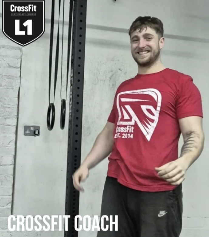 Coach Tom smiling whilst wearing a red CrossFit t-shirt and black Nike shorts standing in a gym with gymnastic rings hanging in the background. The image includes a CrossFit Level 1 certification badge and 'CROSSFIT COACH' text at the bottom.