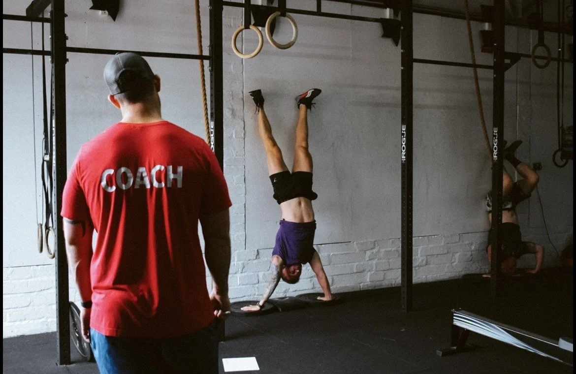 Image of a gymnastics class, with two athletes performing handstand push ups with coach watching.