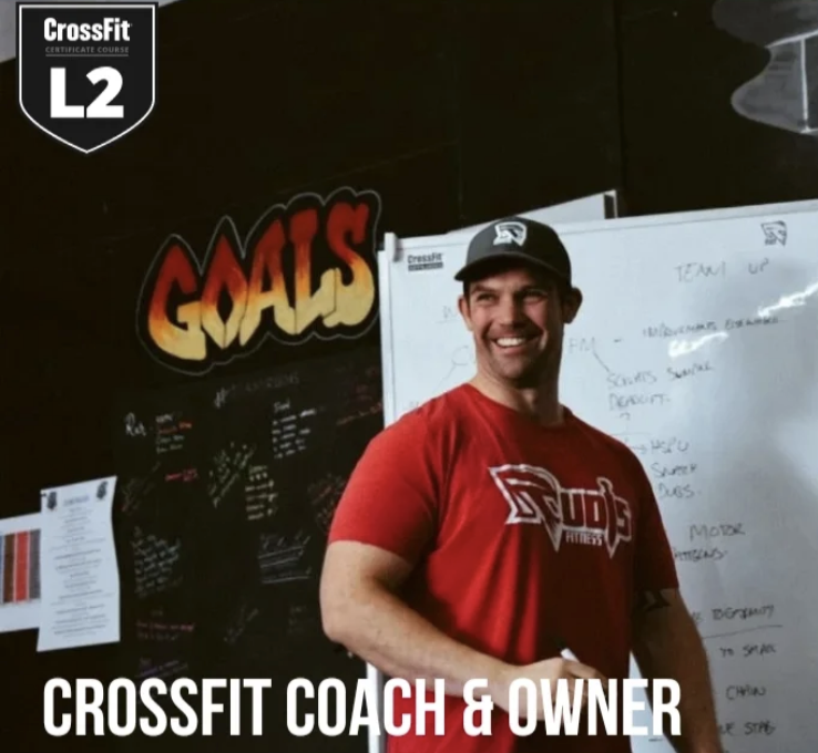 Co-owner and Coach Mitch smiling in his red CrossFit coach T-shirt and black cap posing in front of a whiteboard and a black wall with a large, colourful "GOALS" sign at a CrossFit gym. The image indicates the man is a CrossFit coach and owner.
