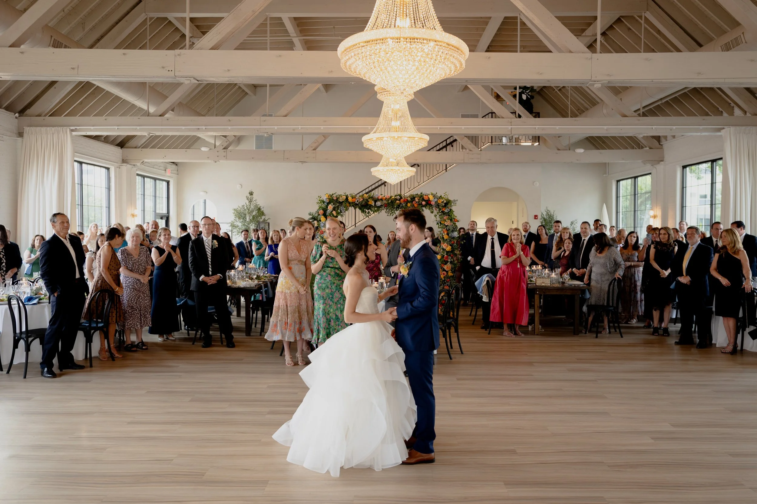 Arbor Loft wedding reception with bride and groom dancing beneath crystal chandeliers in a spacious industrial venue with floral arch and guests celebrating.