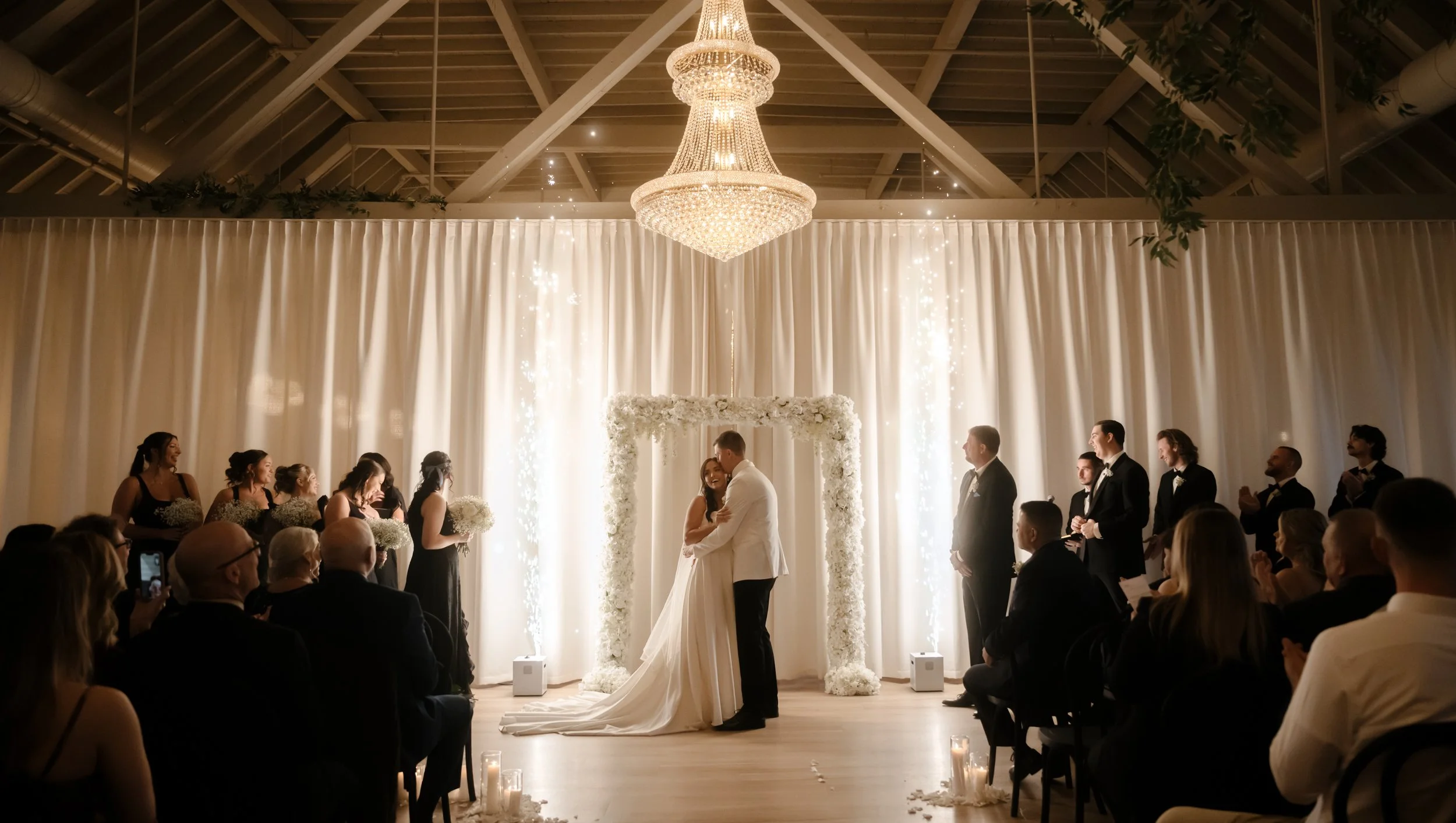 Arbor Loft wedding ceremony with bride and groom under a white floral arch, crystal chandelier, and romantic candlelit aisle in Rochester, NY.