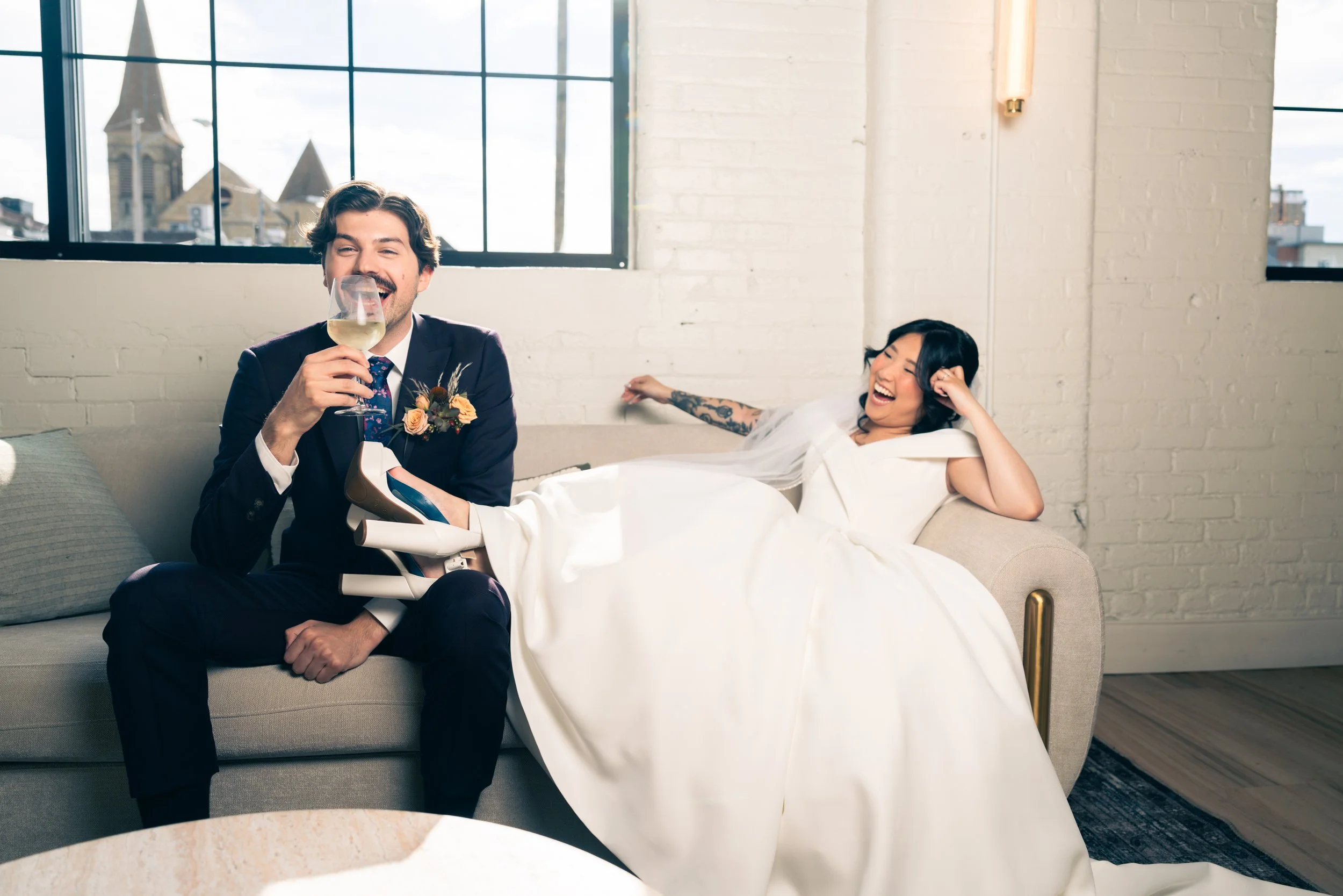 Arbor Loft bride and groom laughing and relaxing on a modern lounge sofa with champagne in a stylish industrial wedding venue.