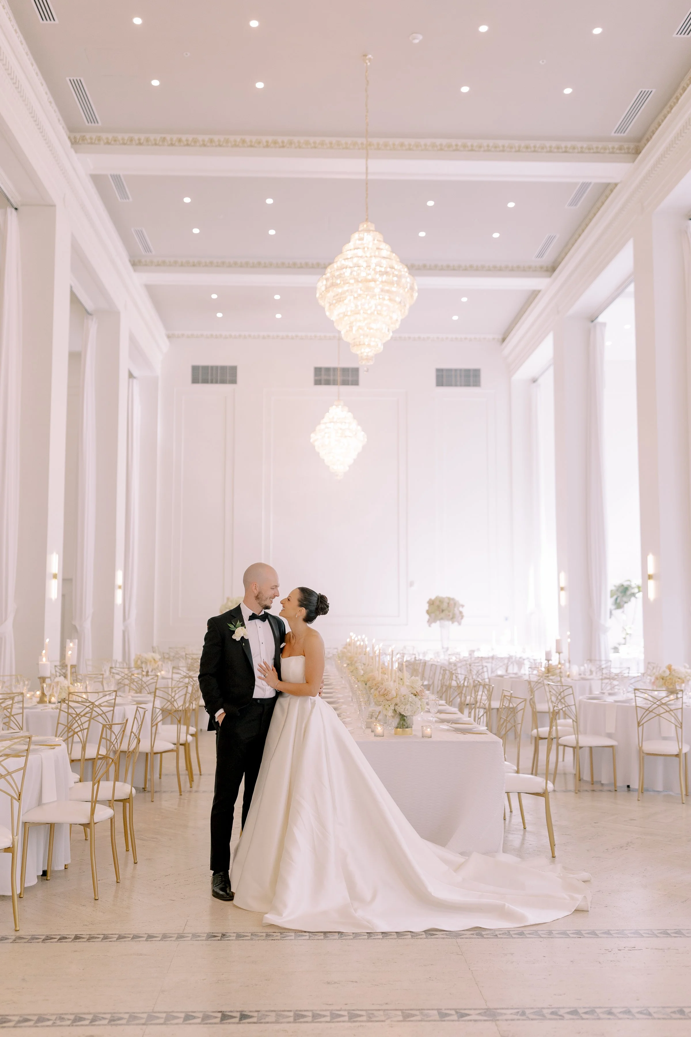 Bride and groom standing in Arbor Midtown’s bright modern ballroom with elegant reception tables, gold chairs, and crystal chandelier in Rochester, NY.