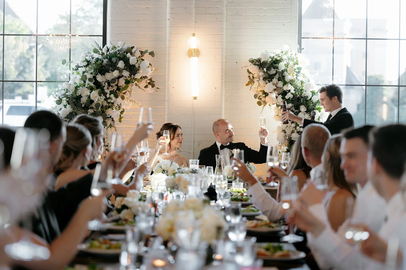 Arbor Loft wedding reception toast with bride and groom, elegant floral arrangements, and guests celebrating in a modern industrial venue.