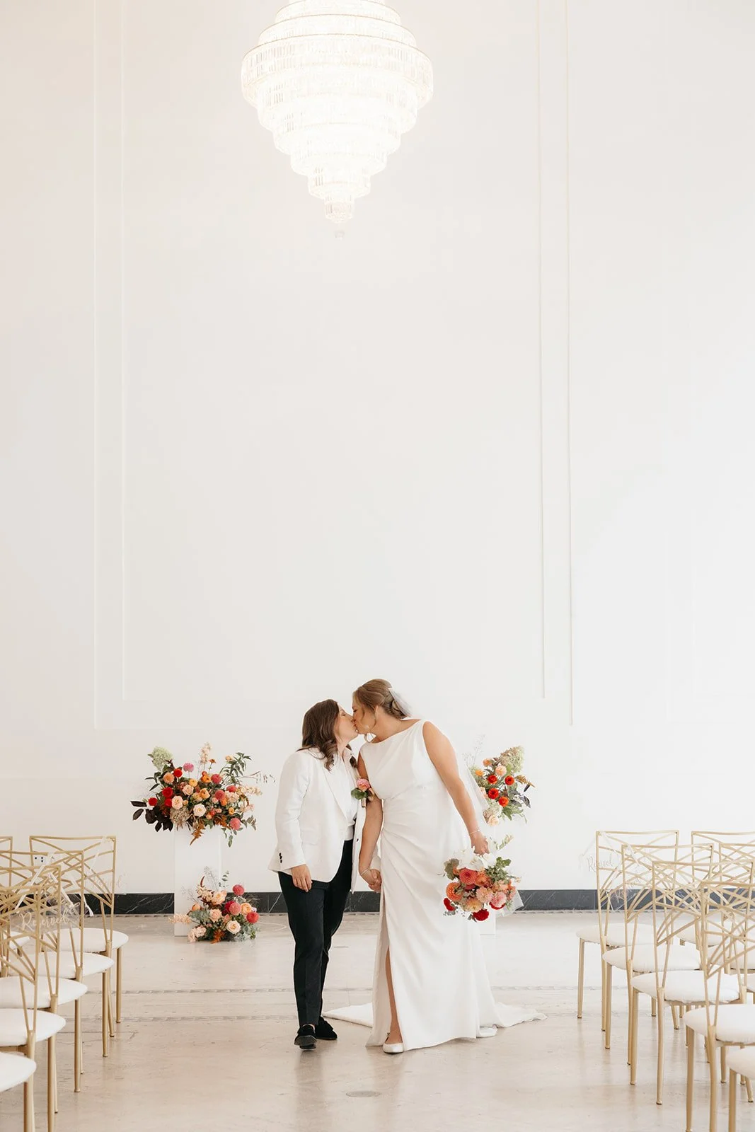 Couple kissing in Arbor Midtown’s bright ceremony space with colorful floral arrangements and gold chairs in Rochester, NY.
