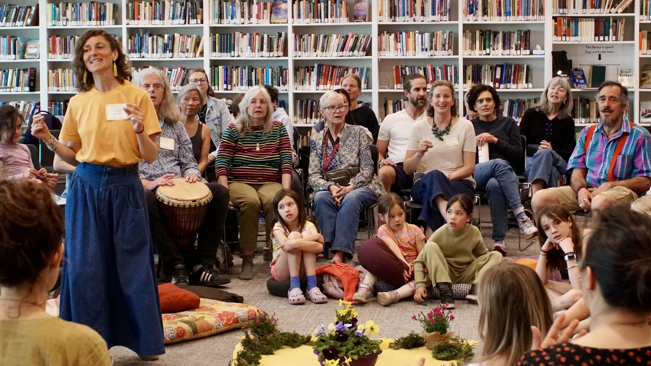 A diverse group of children and adults seated on the floor and chairs in a library, listening to a woman in a yellow shirt with yellow earrings who is standing and speaking, with bookshelves filled with books in the background.