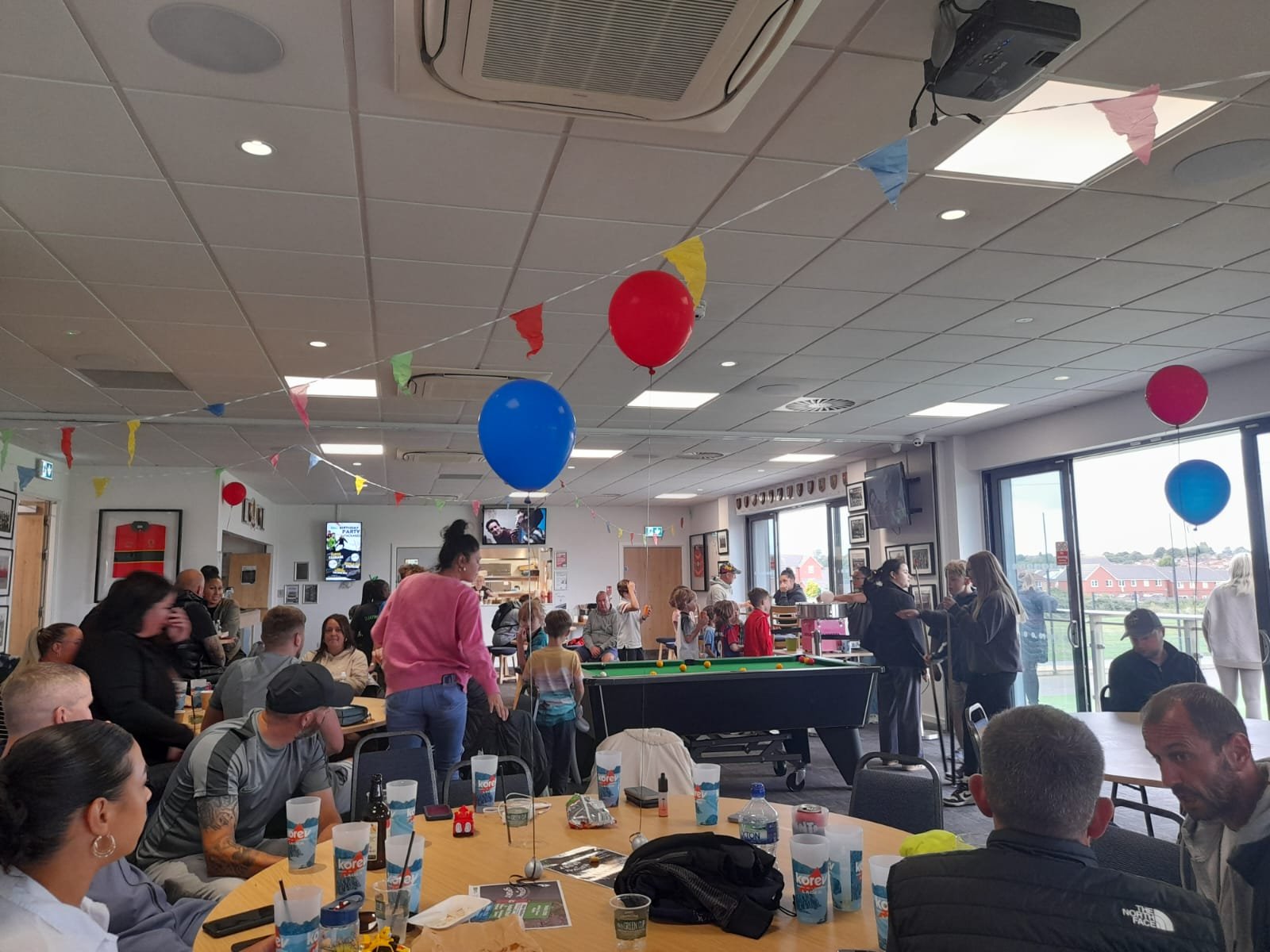 picture of lots of people chatting around tables in Lockleaze Sports Centre. There are balloons and decorations.