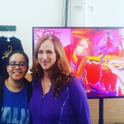 KatMoon Landes stands with another woman at an exhibition of The Broken Hallelujah, with a vivid pink and orange artwork displayed behind them.