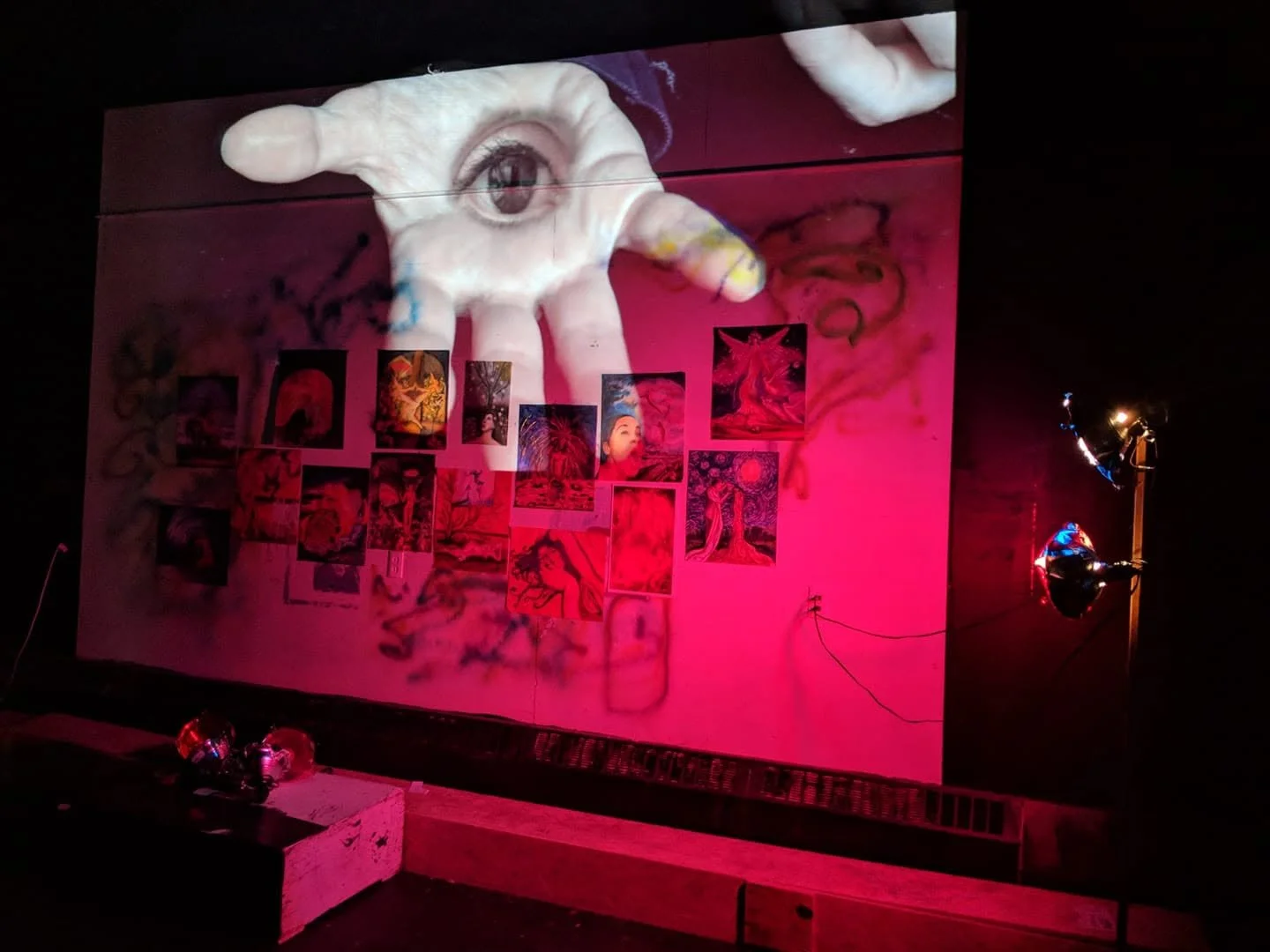 nstallation view of The Witness from The Broken Hallelujah, showing a large projected eye held in an open hand above layered drawings on a red-lit wall.