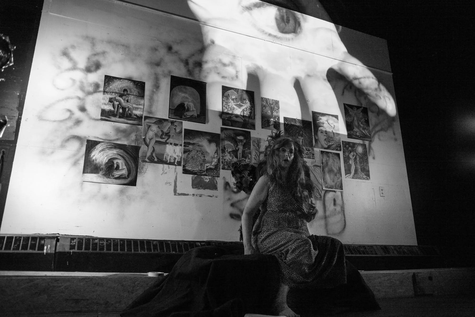Black-and-white installation view of The Broken Hallelujah showing a seated performer beneath a large projection of layered drawings, faces, and an eye.