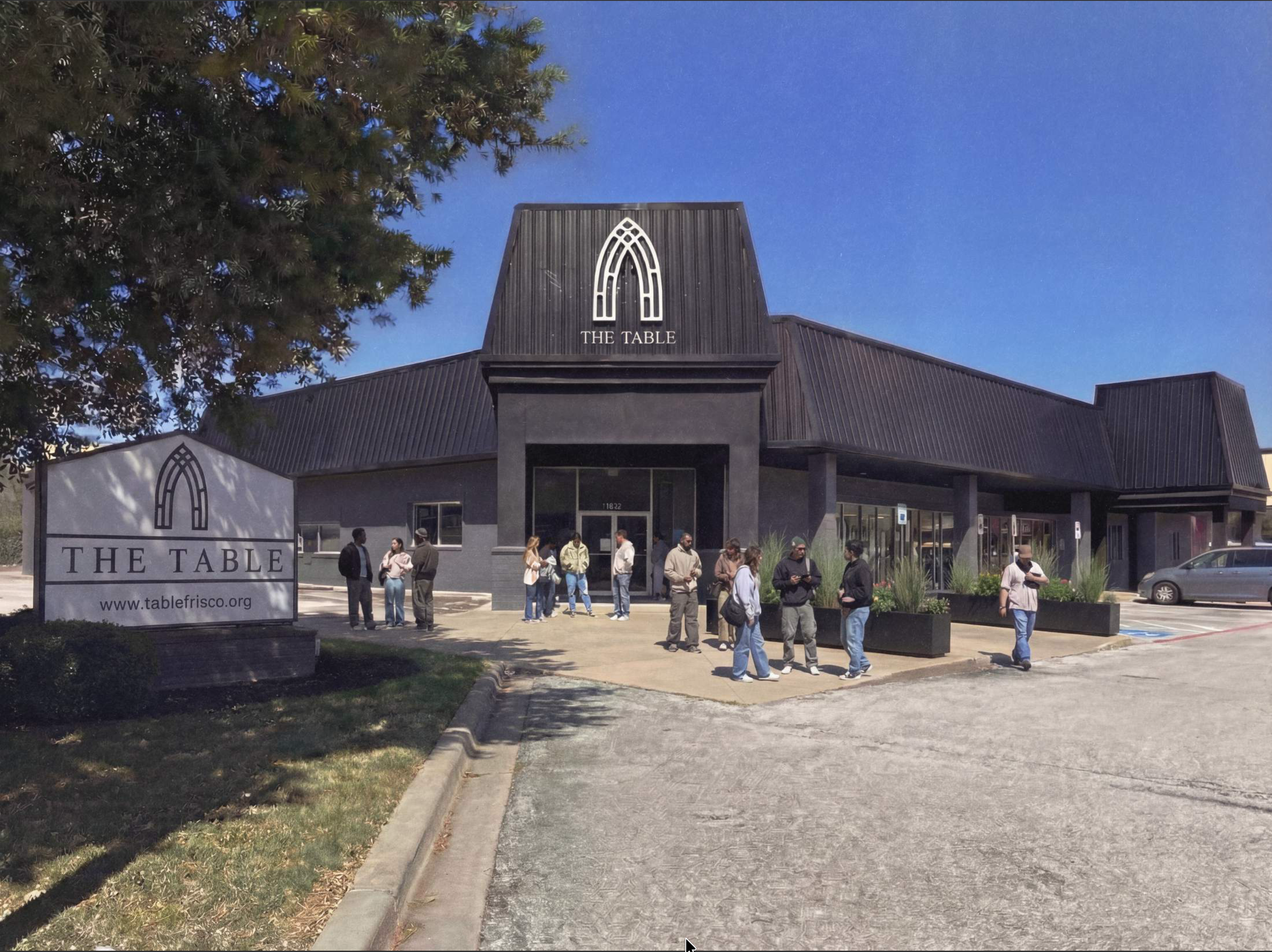 A group of people gathered outside a black building with a sign that reads 'THE TABLE' and an arch-shaped logo above the entrance. The building has a dark metal roof and large windows. A large tree is on the left side of the image, and there is a sign in front of the building displaying the same logo and website 'www.tablefrisco.org'. The sky is clear, and people are walking and standing in front of the entrance.