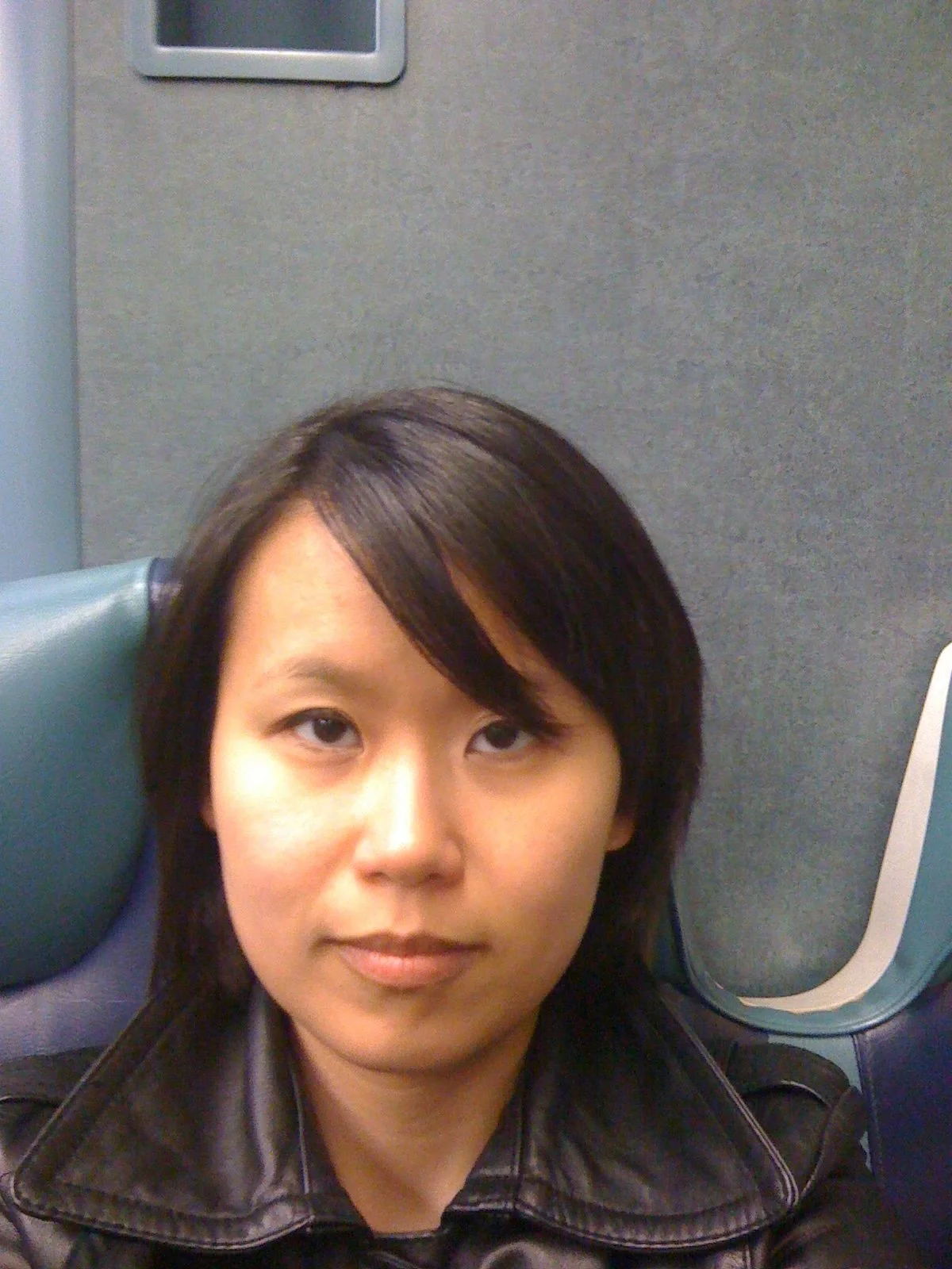 A close-up portrait of Jamie Lee in 2010, featuring a neutral, serious expression and a side-swept fringe. She is wearing a black leather jacket with a prominent collar, seated against the blue and grey interior of what appears to be a train.