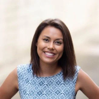Photo of a smiling woman in a blue printed dress. She's looking straight into the camera in a confident smile.