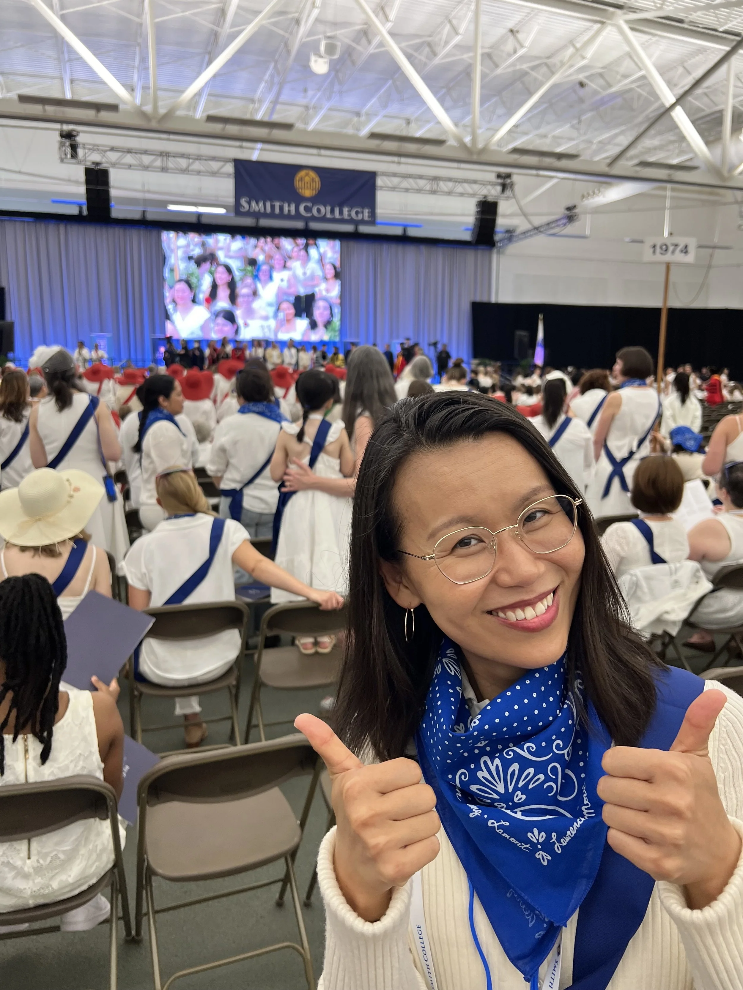 A smiling Jamie Lee with glasses and a blue bandana gives two thumbs up at a Smith College reunion. She is seated in a large indoor arena filled with alumnae wearing white outfits with blue or red sashes.
