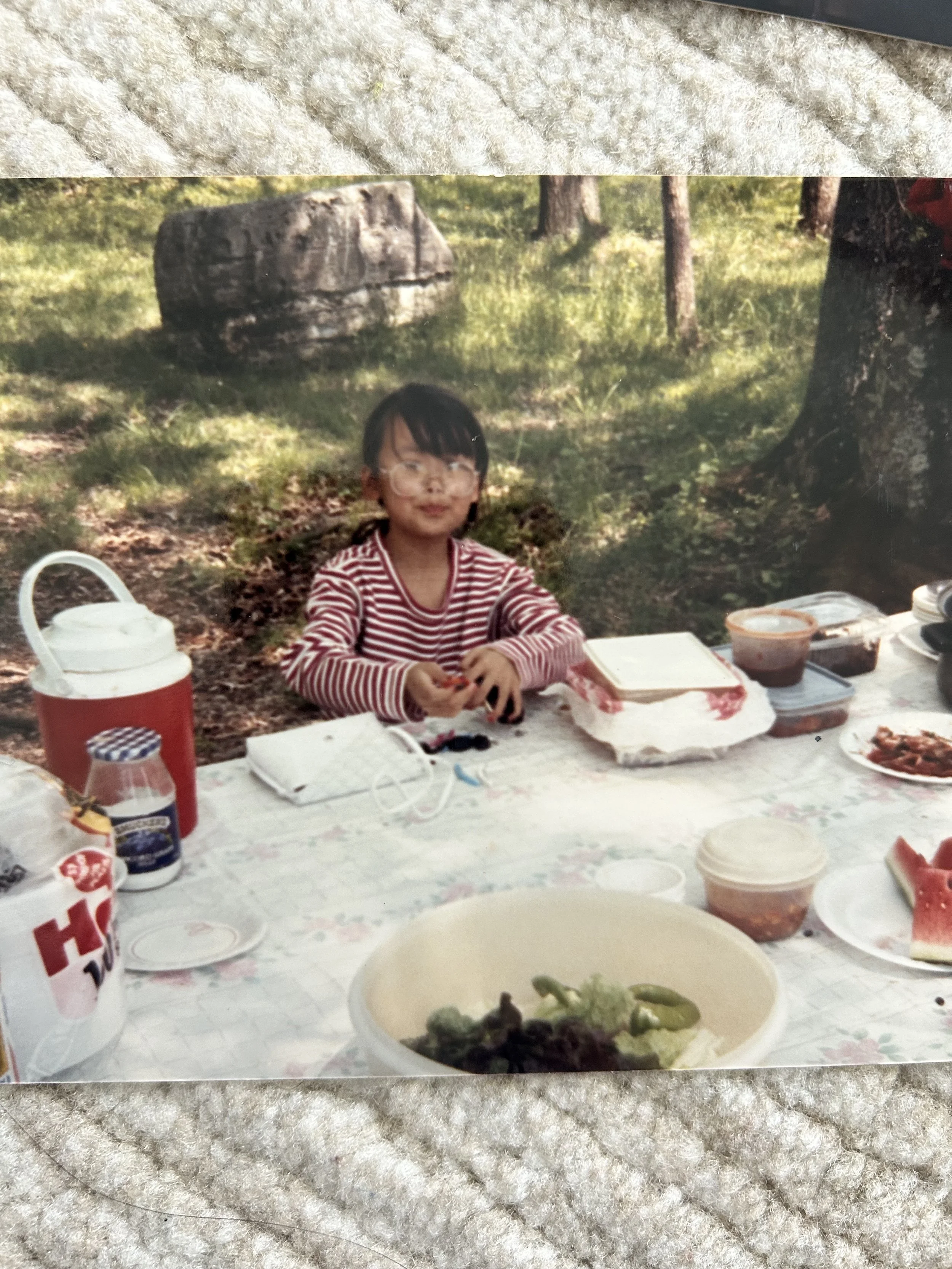 A nostalgic, slightly faded photograph of a young Jamie Lee as a child, sitting at an outdoor picnic table in a wooded area. Jamie wears oversized glasses and a red-and-white striped long-sleeve shirt, looking toward the camera with a subtle smile.
