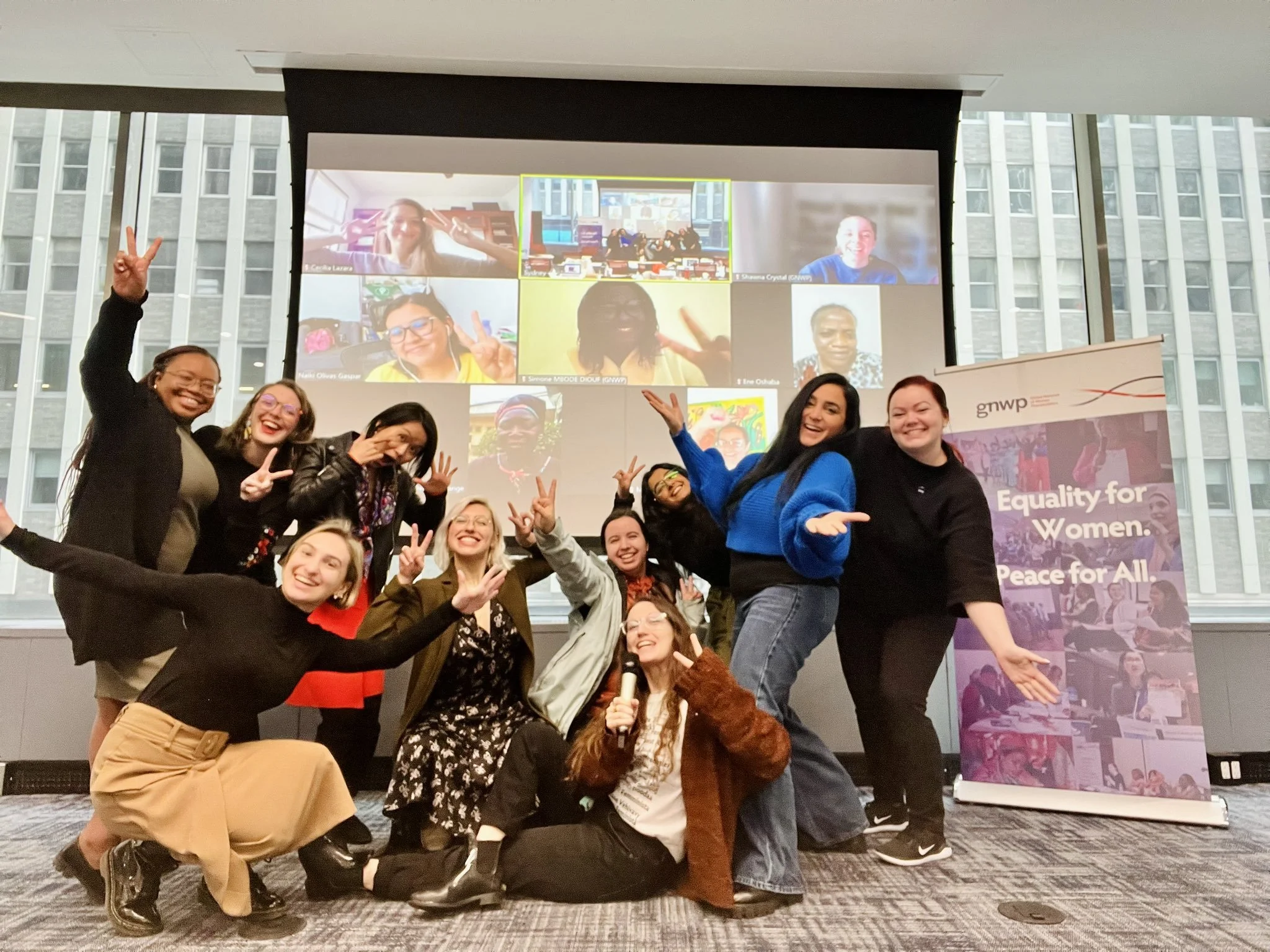 A joyful group of women pose together in a bright, modern office space during a 2023 GNWP Speakers Workshop. They are making peace signs and cheering, with some kneeling in front and others standing.