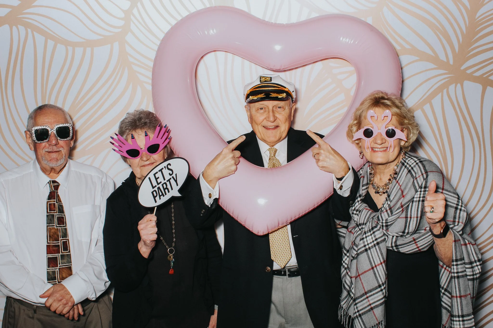Four elderly people at a party, with a man in the center pointing at his head through a large pink heart-shaped balloon. The group is dressed in semi-formal clothing, wearing humorous sunglasses and holding a speech bubble sign that says 'LET'S PARTY