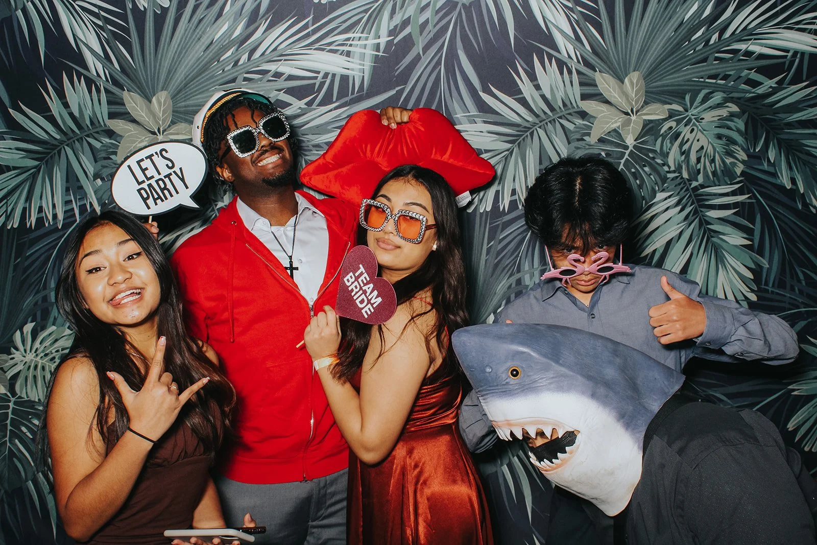 Group of five friends at a party wearing fun costumes and accessories, standing in front of a tropical-themed backdrop with palm leaves. One person is holding a sign that says 'Let's Party,' another has a heart-shaped sign that says 'Team Bride,' one