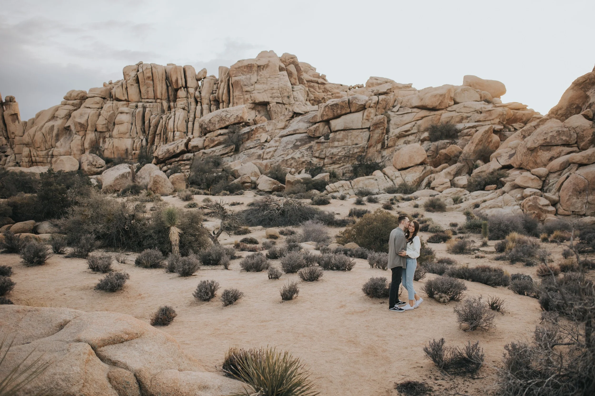 A couple standing in a desert landscape with large rock formations and sparse desert vegetation at joshua tree national park
