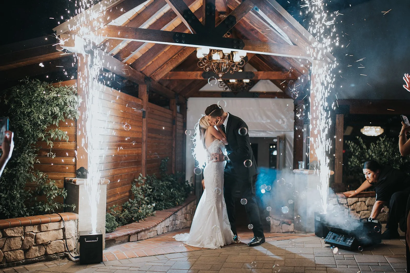 A bride and groom share a kiss beneath fireworks at their wedding reception in a wooden pavilion with a rustic ambiance at wedding venue in temecula named los willows