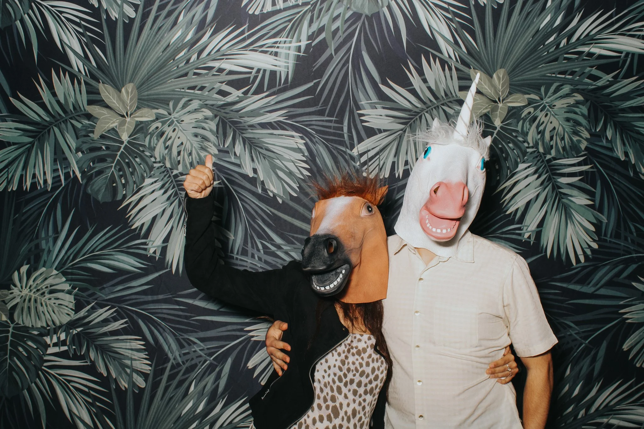 Two people wearing horse and unicorn masks, standing in front of a tropical leaf patterned background. One person is giving a thumbs-up.