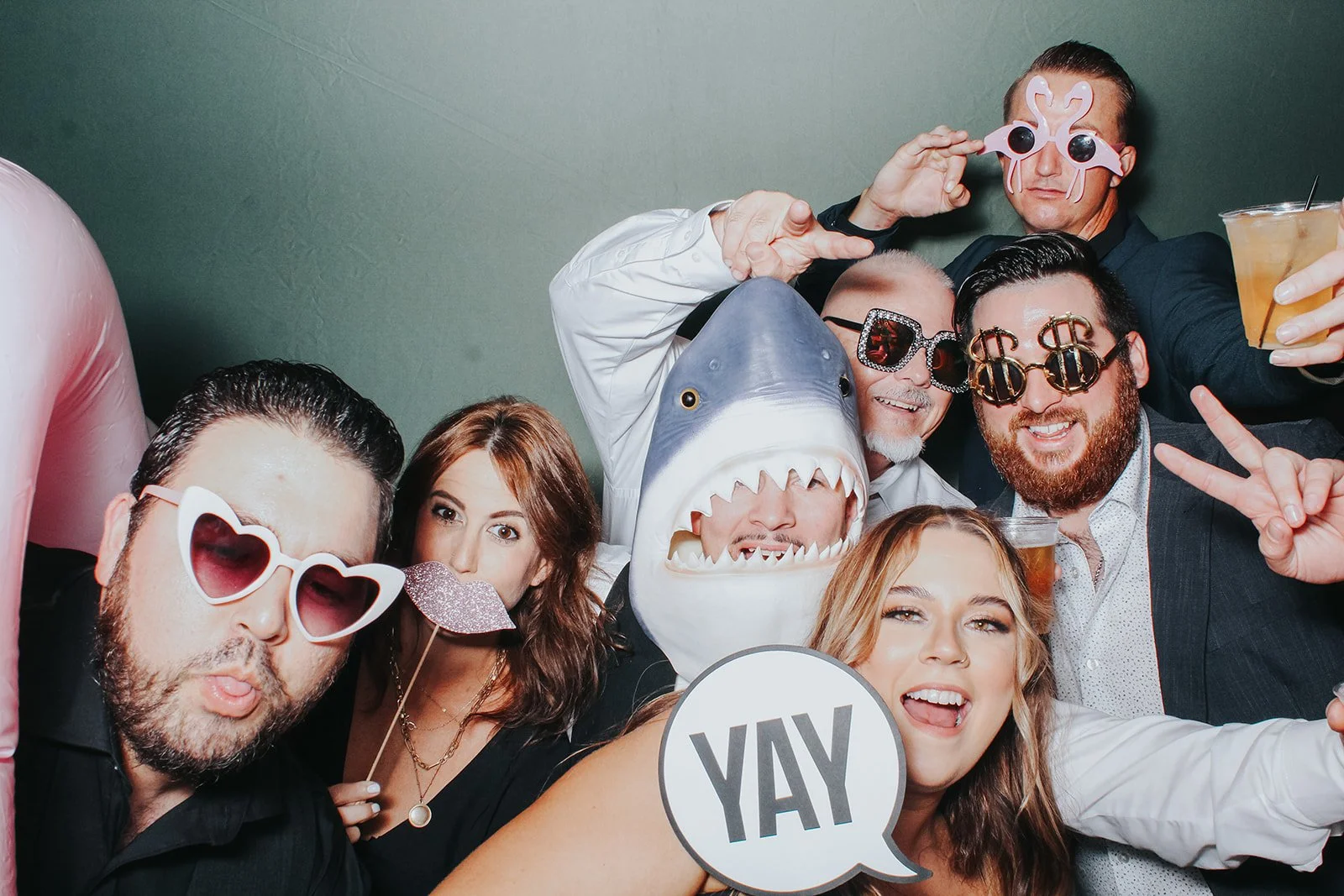 Group of people at a party wearing fun glasses and costumes, holding drinks, with a person in a shark costume, a person with a pink mask, and a woman holding a 