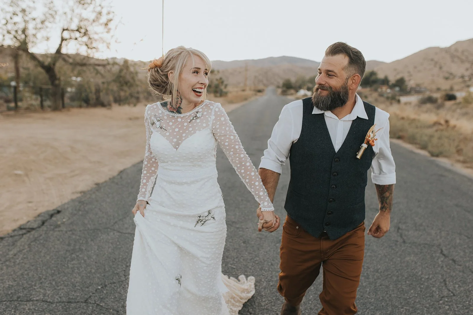 A happy couple holding hands and walking on a rural road in a desert landscape, dressed in wedding attire.