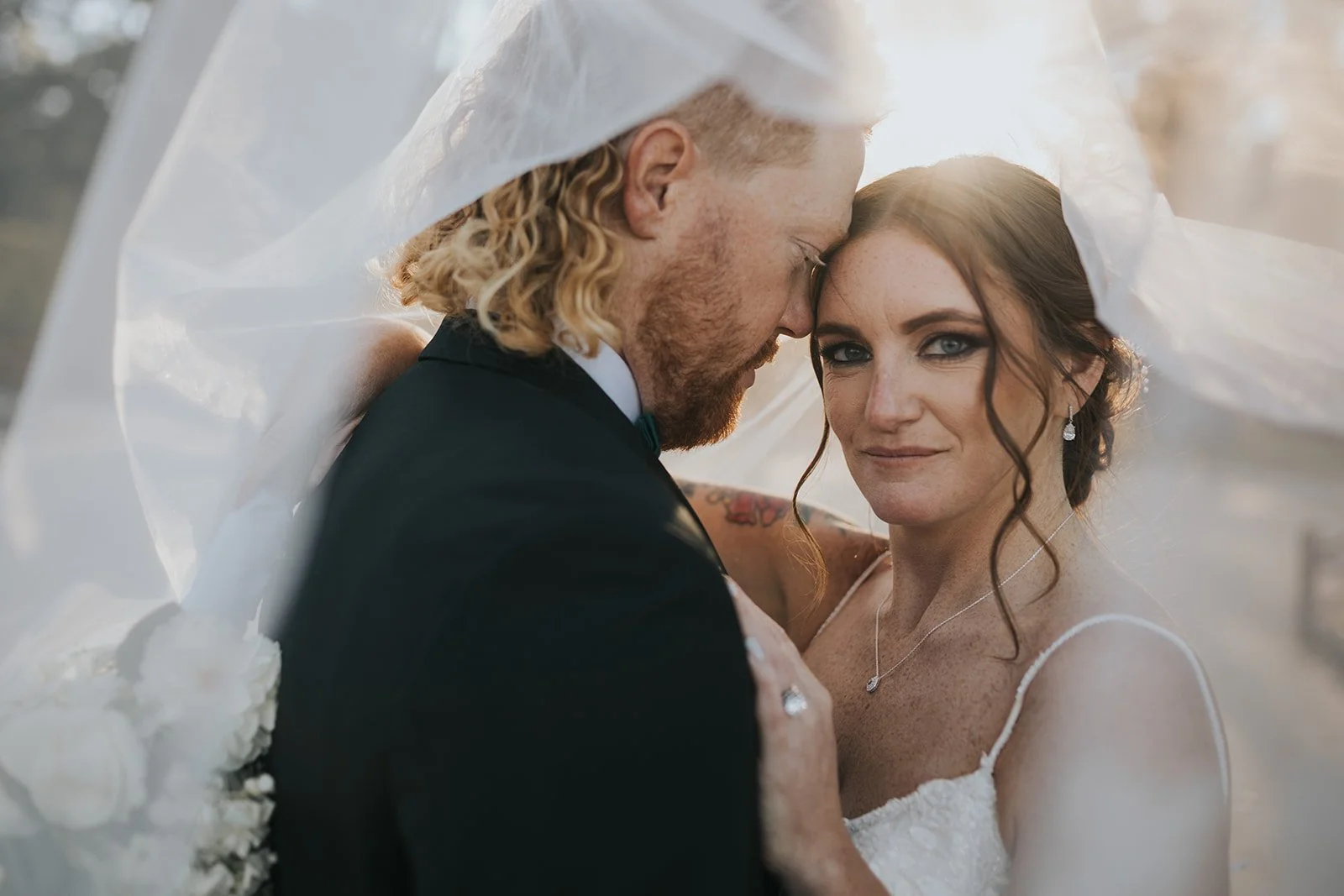 A bride and groom close together, with their foreheads touching, under a veil during their wedding photoshoot outside.