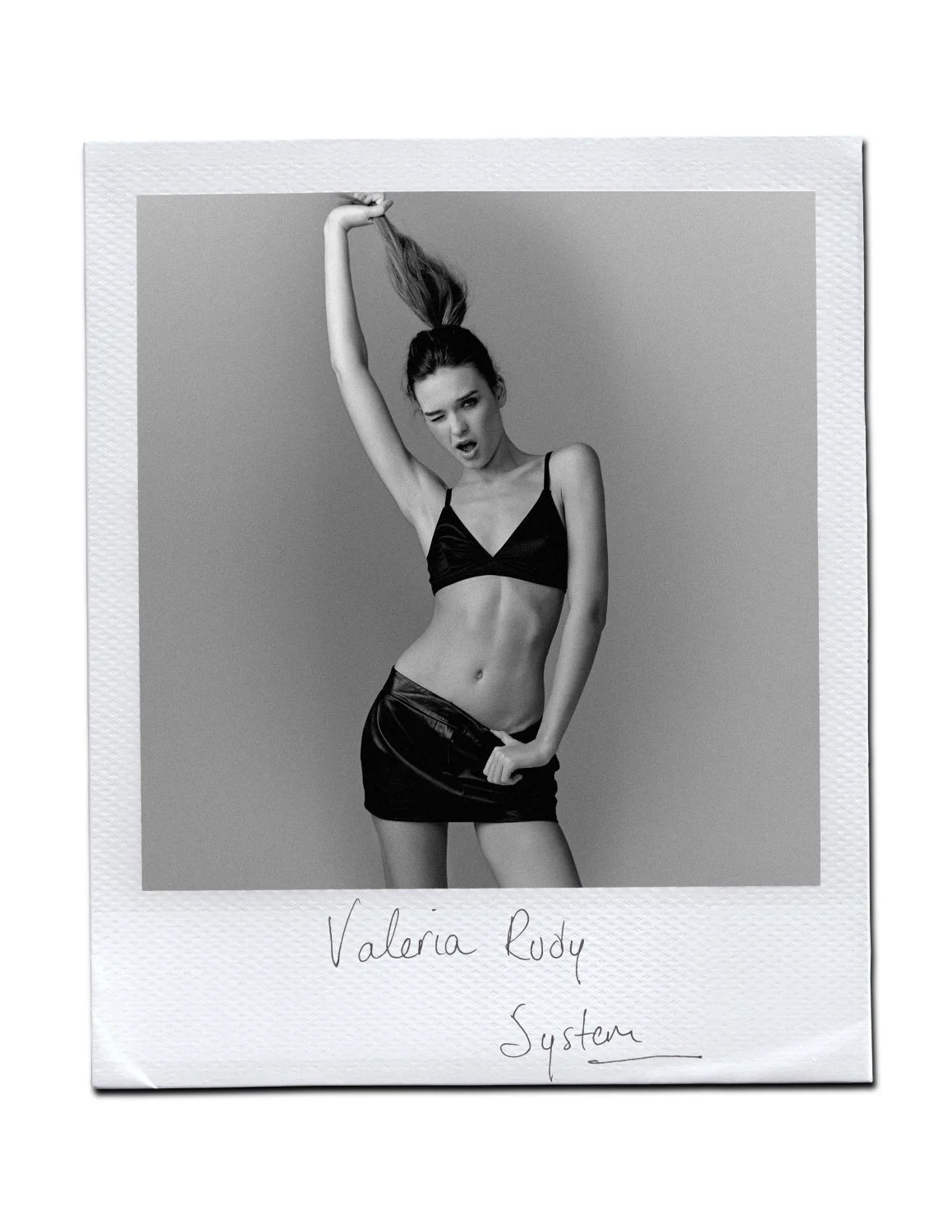 Person posing in black outfit, holding hair up, monochrome photo.