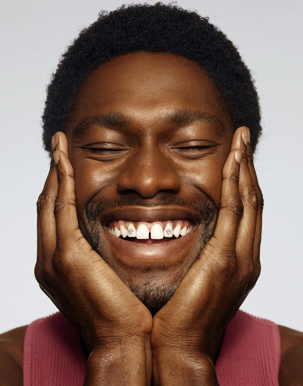 Man smiling widely with hands on cheeks, wearing maroon top, showing diamond-shaped tooth gems.
