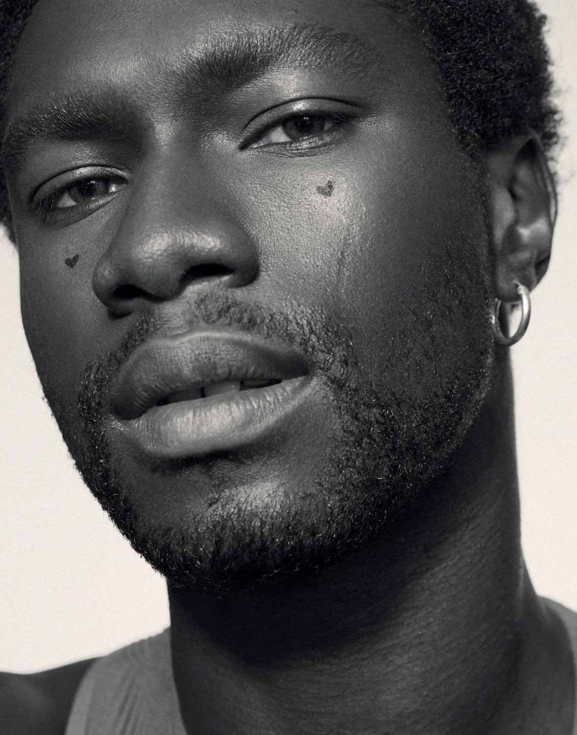 Close-up of a person's face with heart-shaped tattoos near the eyes, wearing a hoop earring.