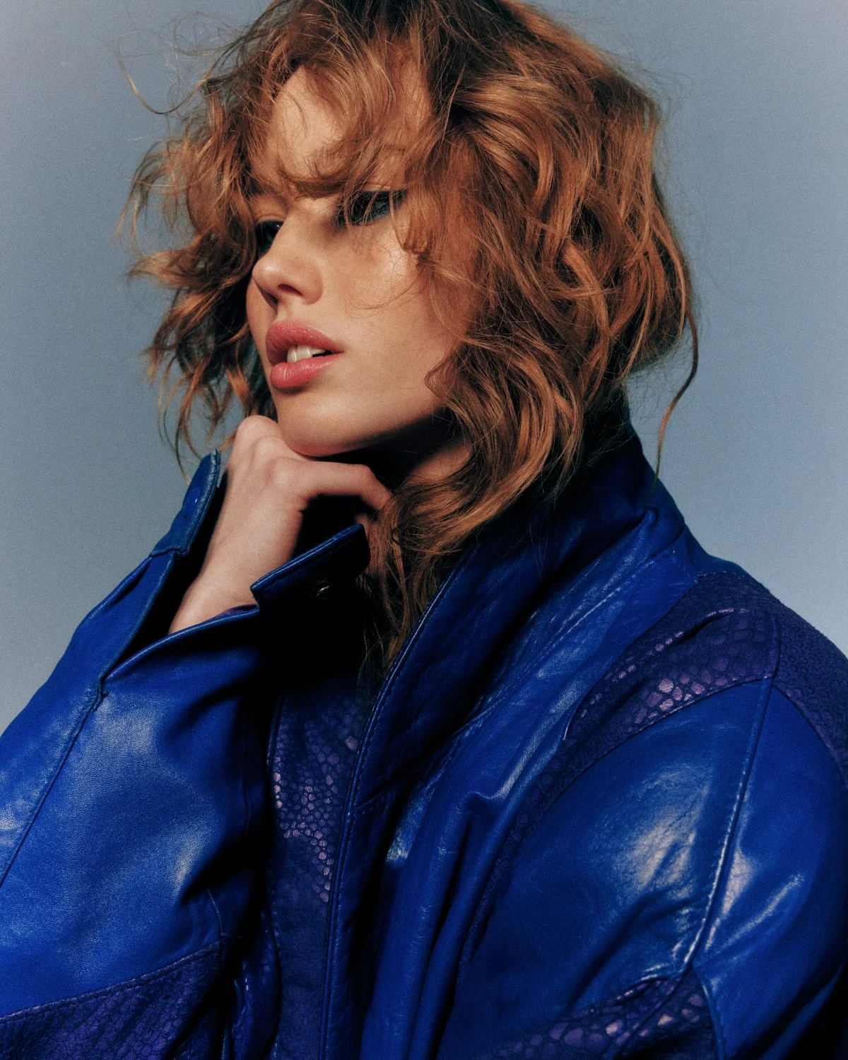 Person wearing a blue leather jacket with tousled, curly hair against a neutral background.