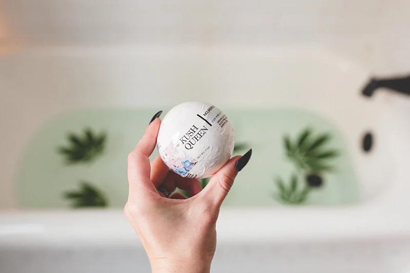 Model holding white Shield 100mg Immunity Bath Bomb in front of tub with clear water and floating hemp leaves. 