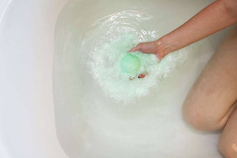 Model kneeling in tub with clear water holding dissolving green Relieve CBD bath bomb by Kush Queen. 