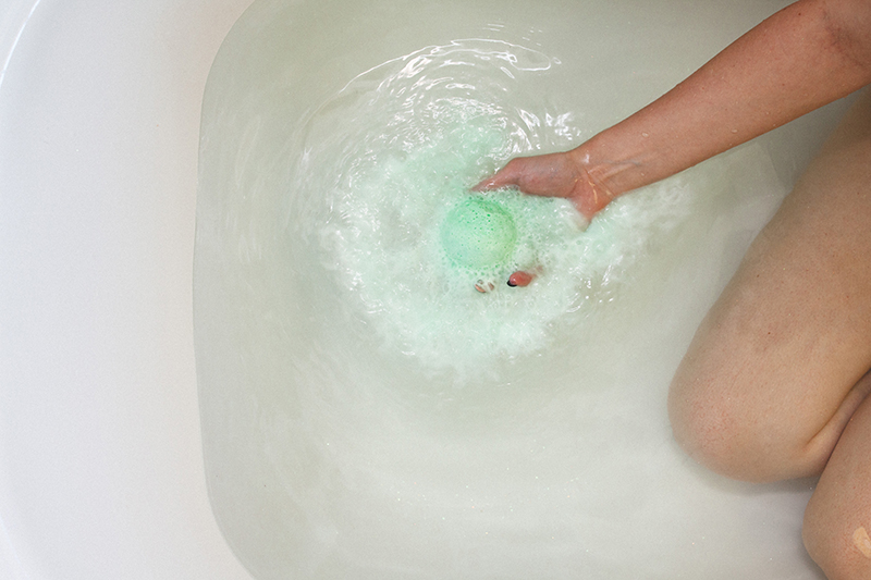 Model kneeling in tub with clear water holding dissolving green Relieve CBD bath bomb by Kush Queen.