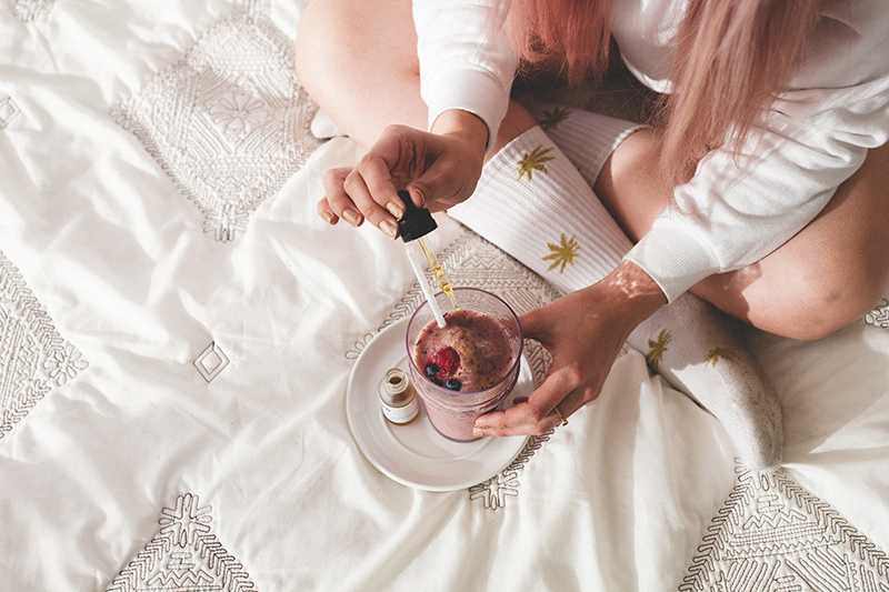 Model with pink hair adding full dropper of Bare tincture to colorful fruit smoothie sitting on bed. 