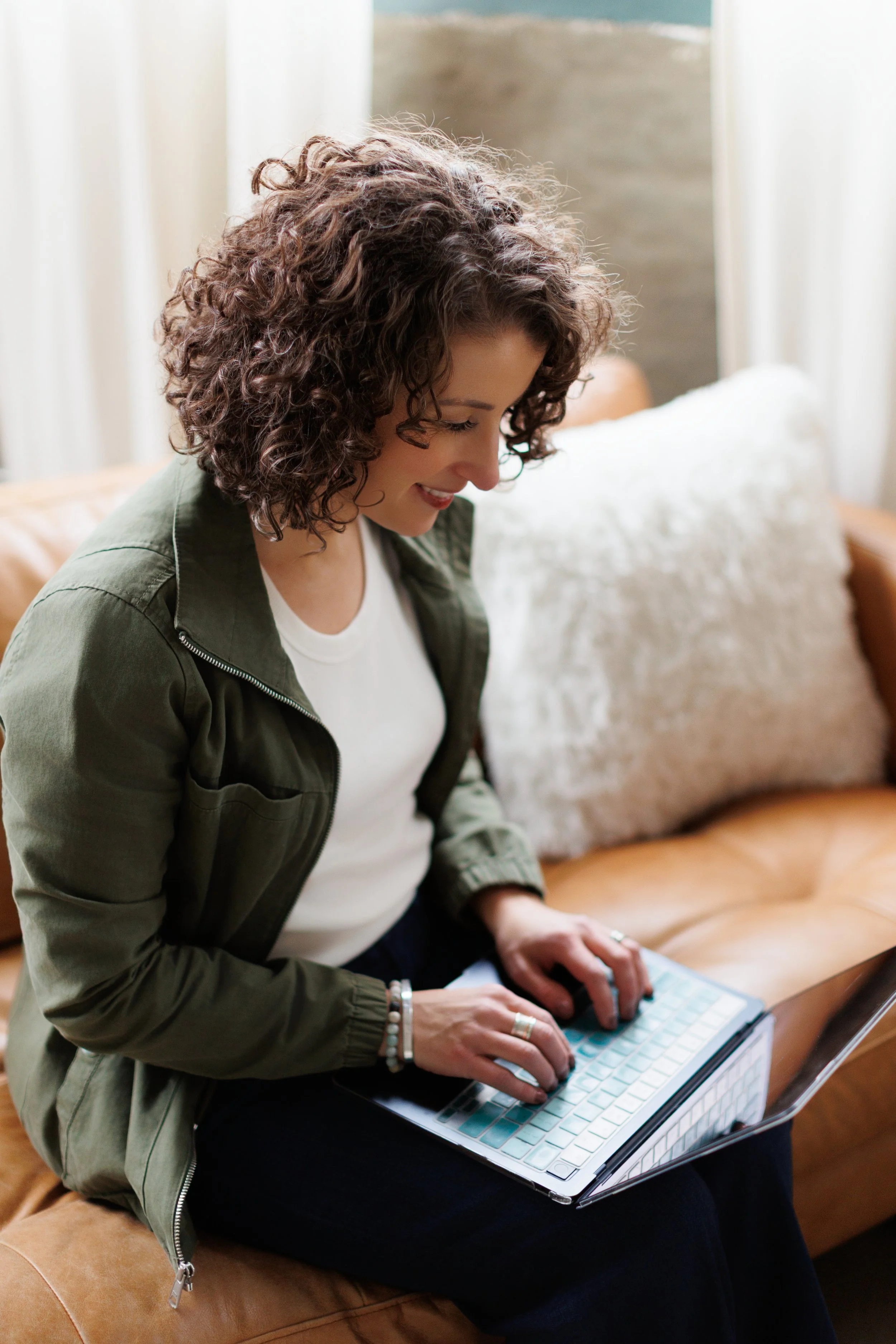 A woman with curly hair wearing a green jacket and white shirt, sitting on a tan leather couch with a pillow, using a laptop.