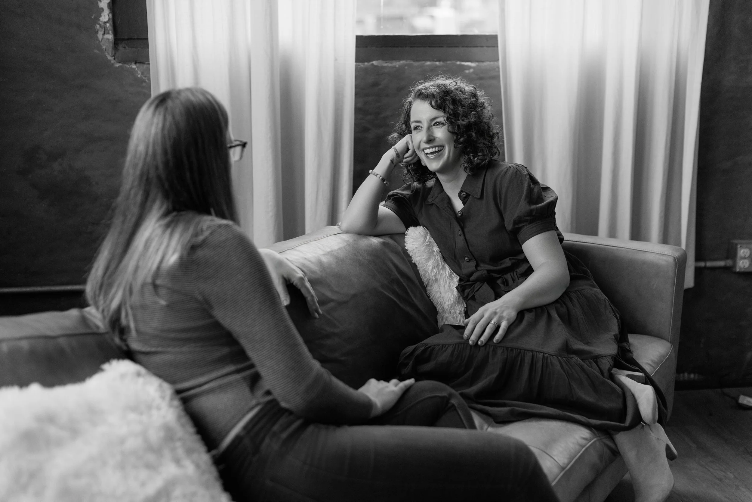 Two women having a conversation on a sofa, laughing and smiling, in a cozy room with curtains and textured walls.