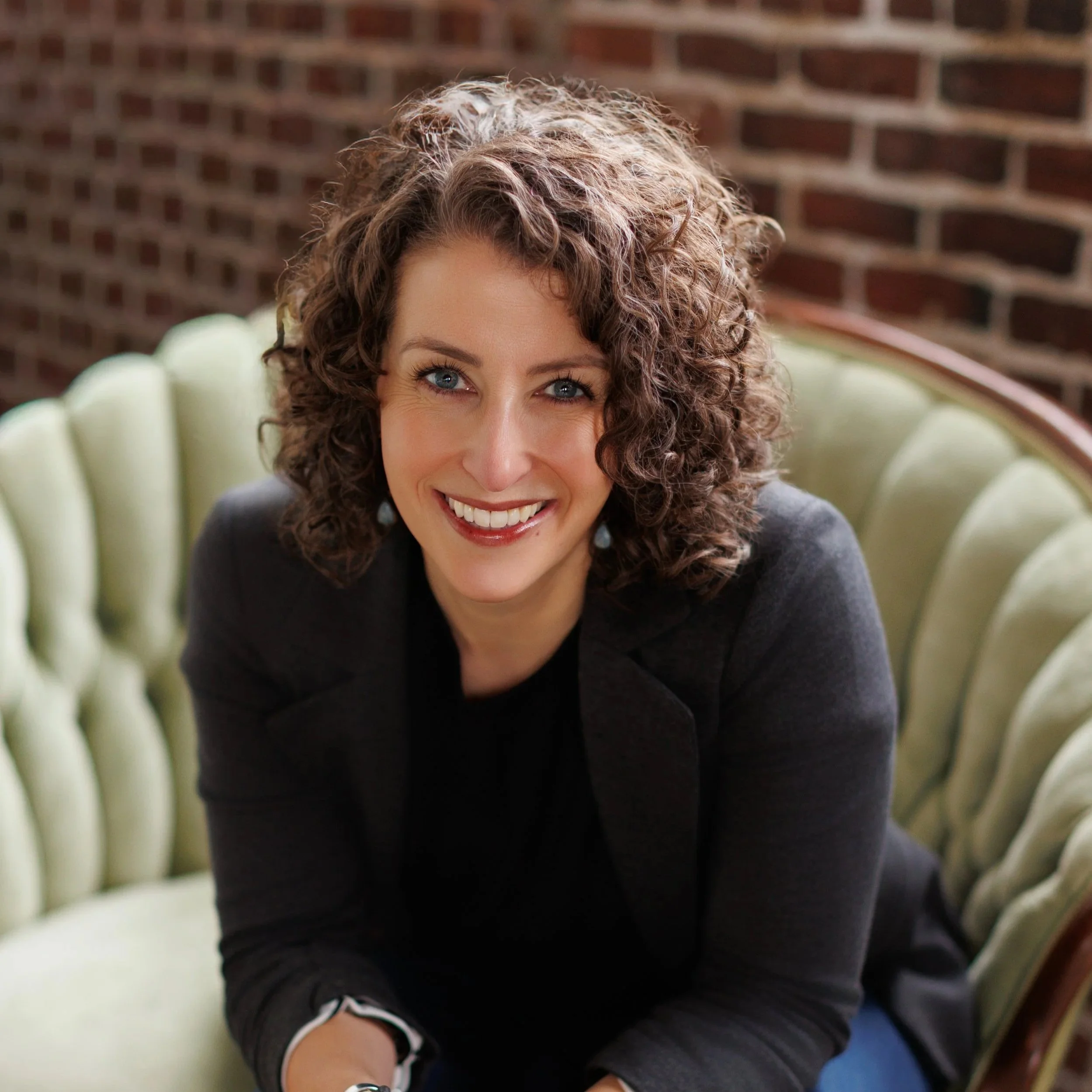 A woman with curly brown hair and blue eyes smiling, sitting on a green vintage sofa with a brick wall in the background.