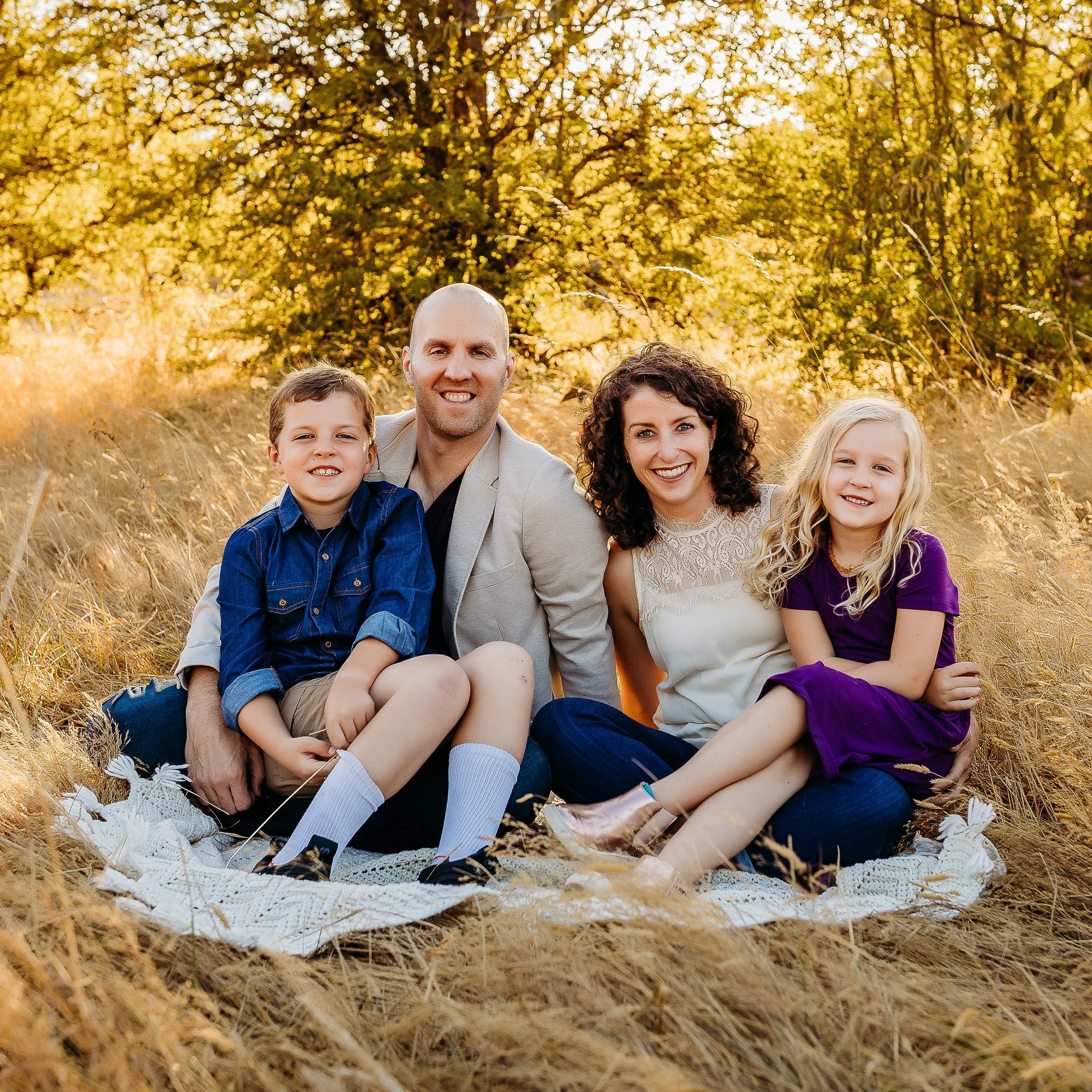Family of four sitting on a blanket in a grassy field during autumn, smiling at the camera.