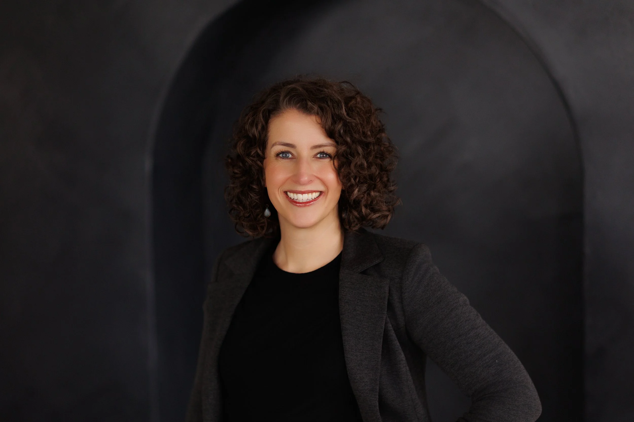 A woman with curly brown hair, smiling, wearing a black top and dark blazer, standing against a dark arched background.
