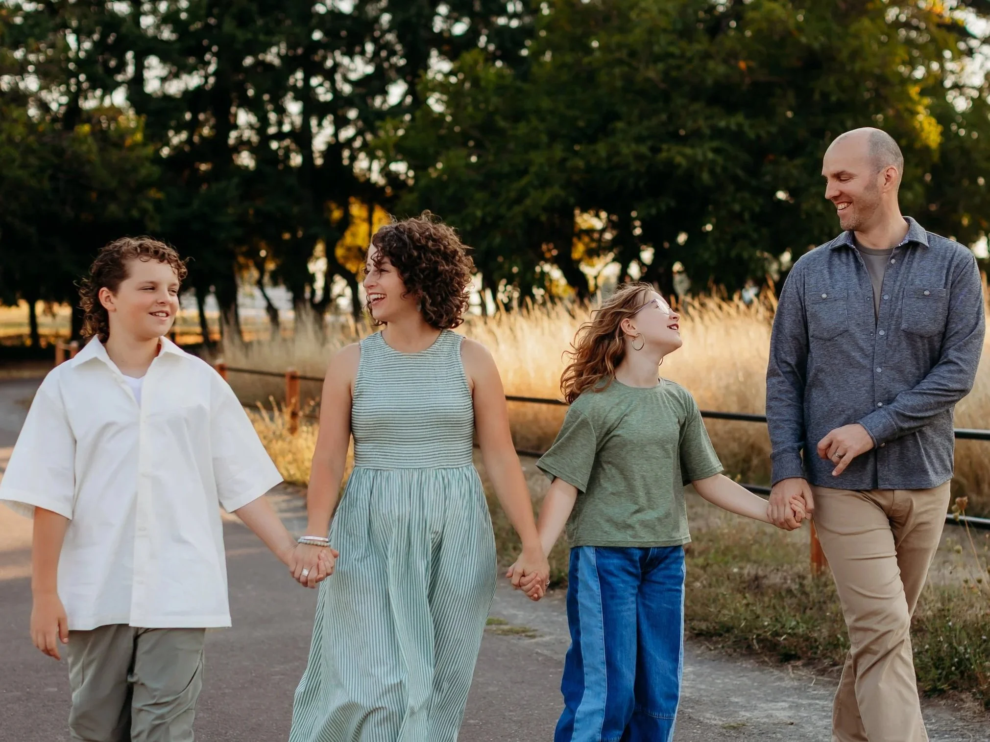 A family of five holding hands and walking outdoors during sunset. The scene is in a park or nature trail with trees and tall grass in the background. They are smiling and looking at each other.