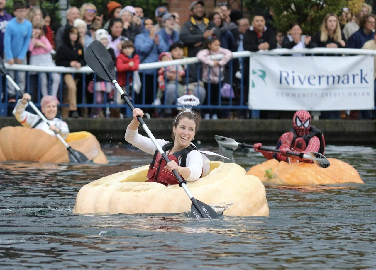 People kayaking in pumpkin-shaped boats during a festive event with spectators watching from the shore.