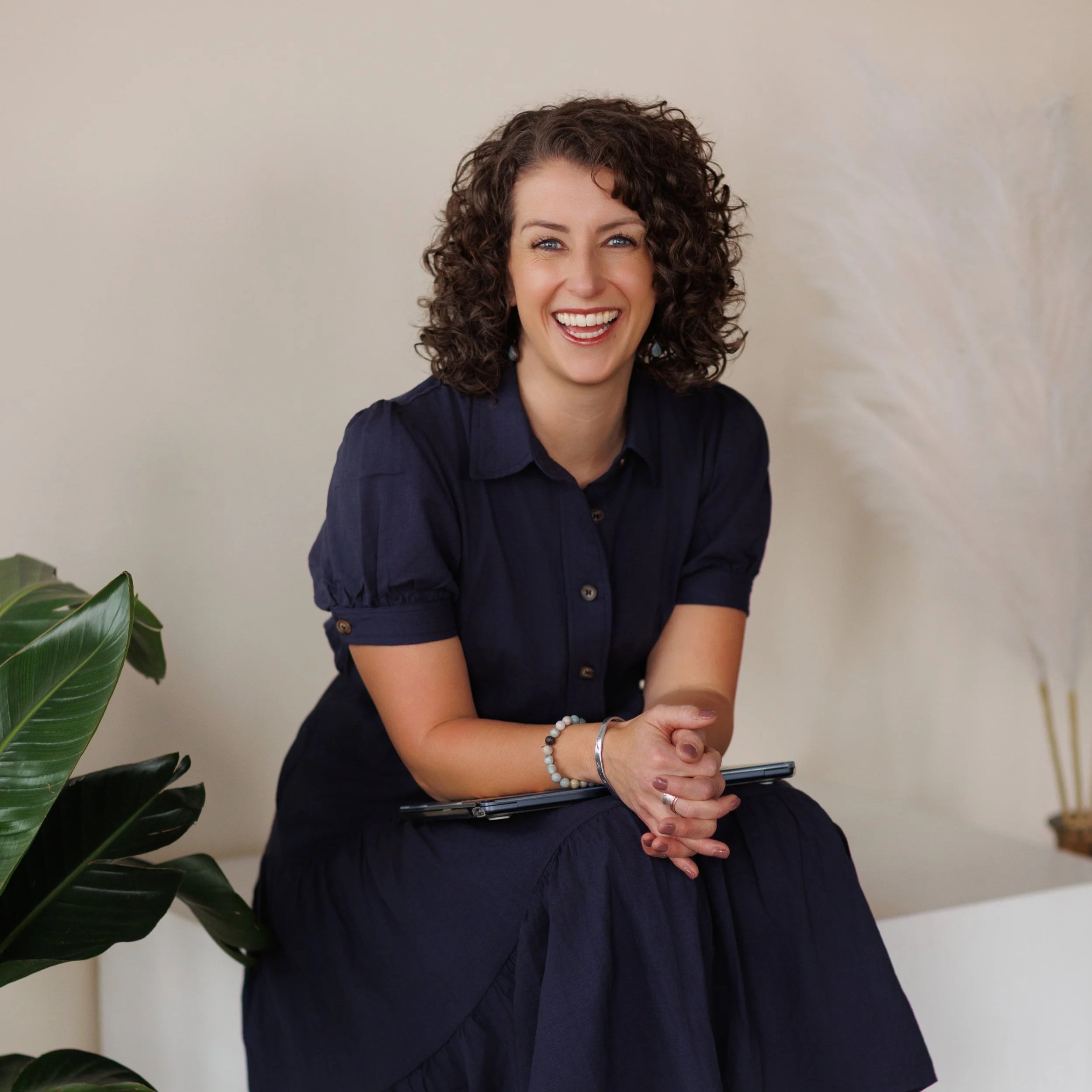 Photo of a woman with curly brown hair wearing a navy blue dress, smiling, sitting on a white couch with greens on the side and a neutral background.
