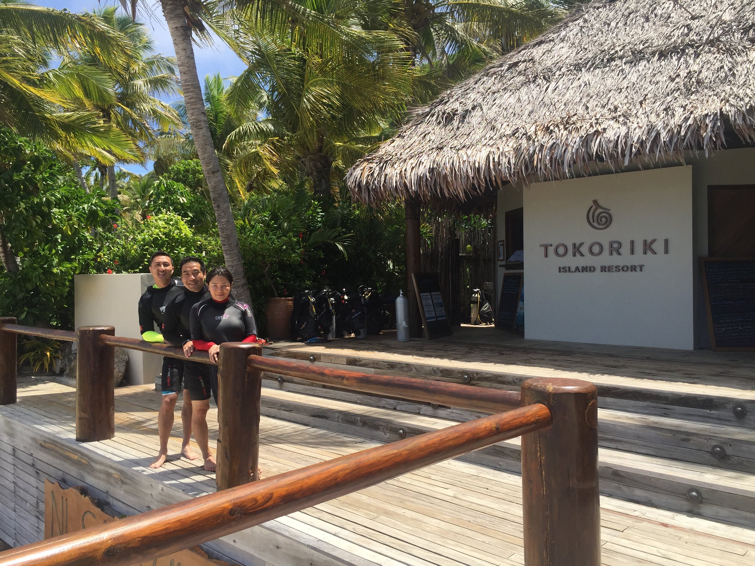 People in swimsuits standing on a wooden deck outside a tropical resort with palm trees and signage that reads 'Tokoriki Island Resort'.