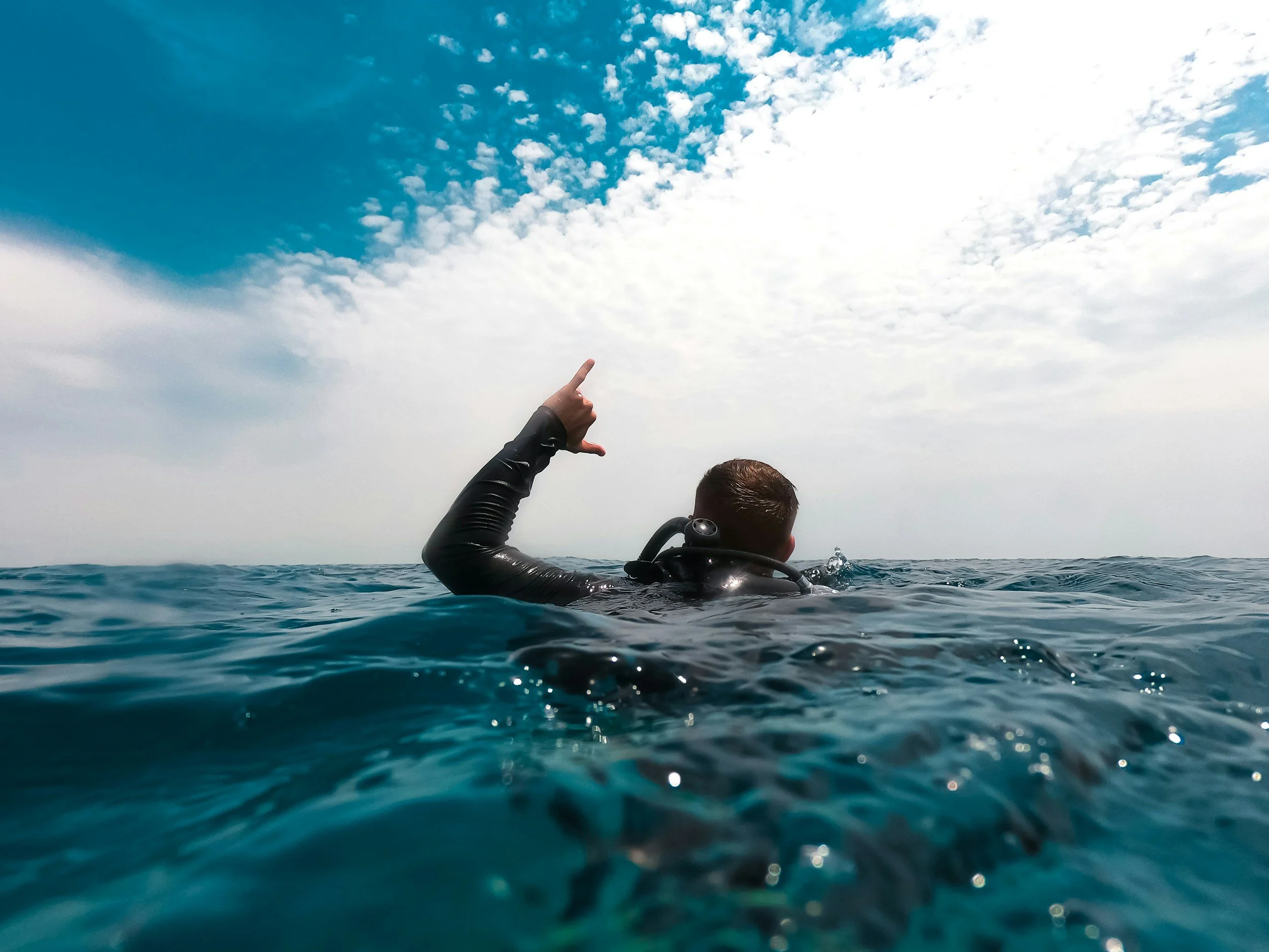 Person wearing a wetsuit in the water, pointing towards the sky with a cloudy sky and sunlight in the background.